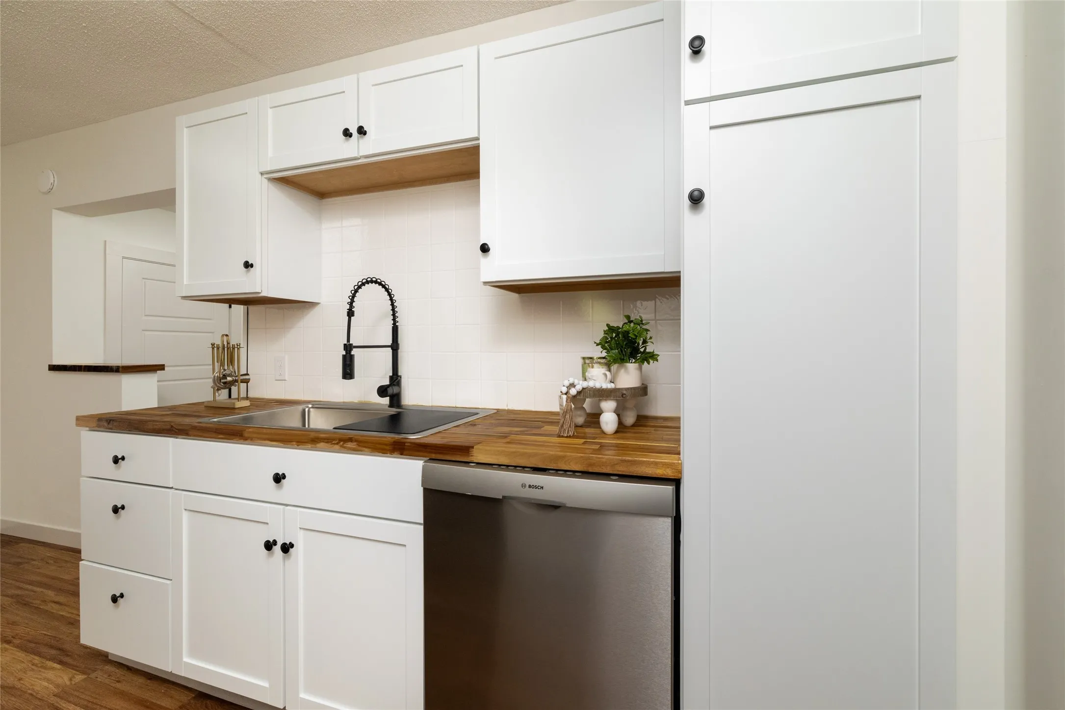 Modern kitchen featuring white shaker cabinetry, wood-finish countertops, a stainless steel double sink with a matte black gooseneck faucet, a white subway tile backsplash, and wood-finish flooring