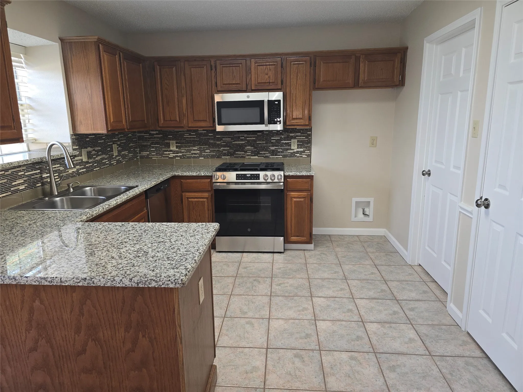 Kitchen featuring wood cabinetry, light-toned granite-style countertops, stainless steel appliances, a mosaic tile backsplash, and ceramic tile flooring