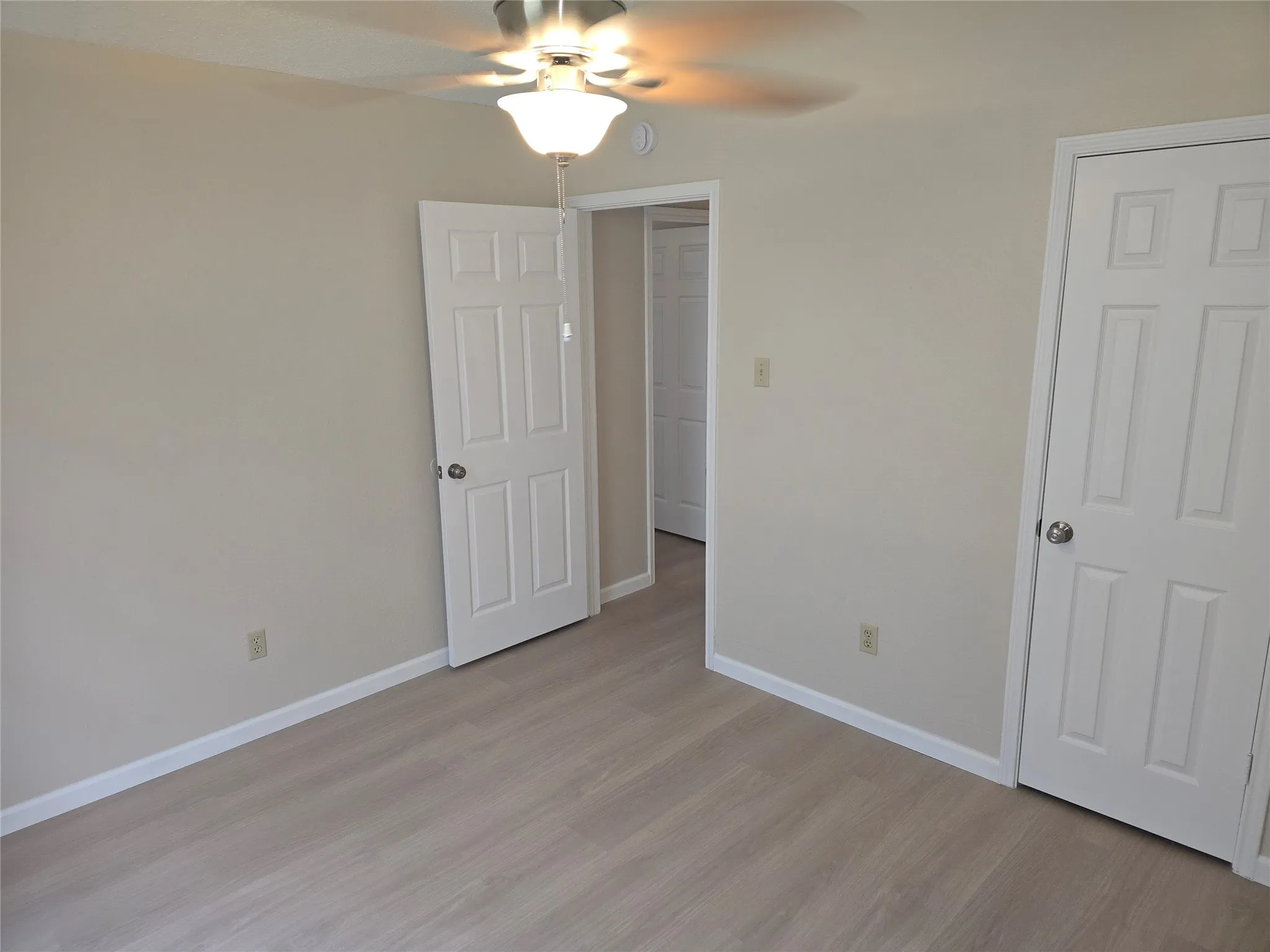 Neutral-toned room featuring light wood-finish flooring, a ceiling fan with integrated lighting, and white six-panel doors