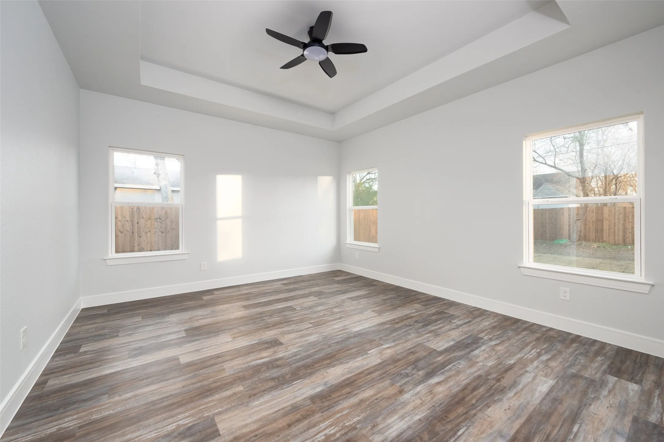 Spacious room featuring wood-finish flooring, white baseboards, and a tray ceiling with a ceiling fan