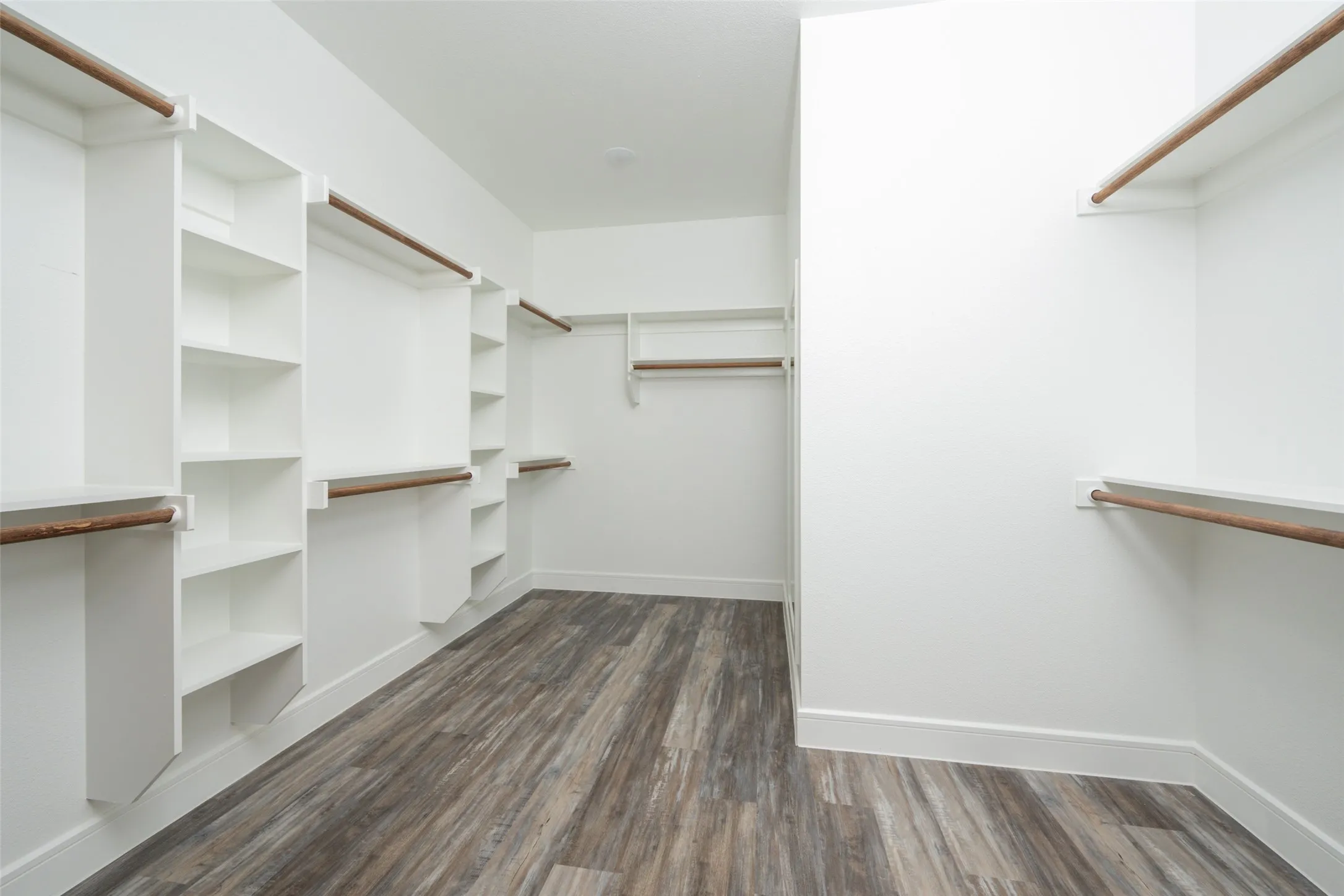 Spacious walk-in closet featuring extensive built-in shelving, multiple hanging rods, clean white walls, and wood-finish flooring