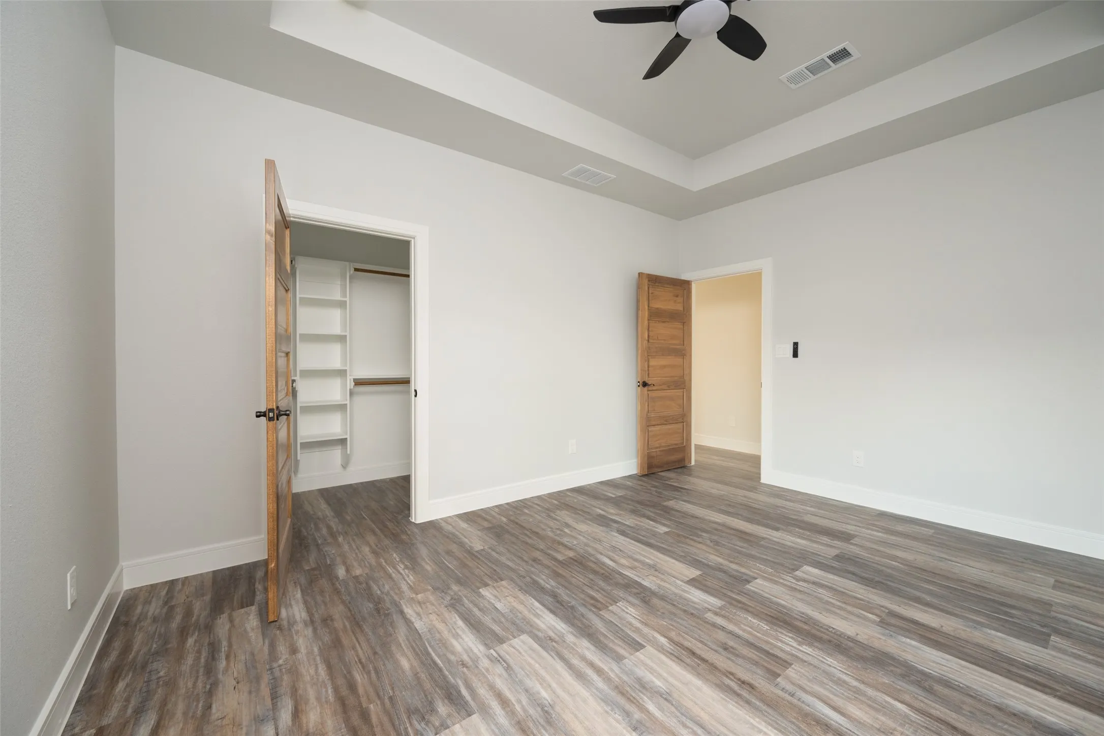 Spacious room featuring wood-finish flooring, light grey walls, and a tray ceiling with a built-in ceiling fan
