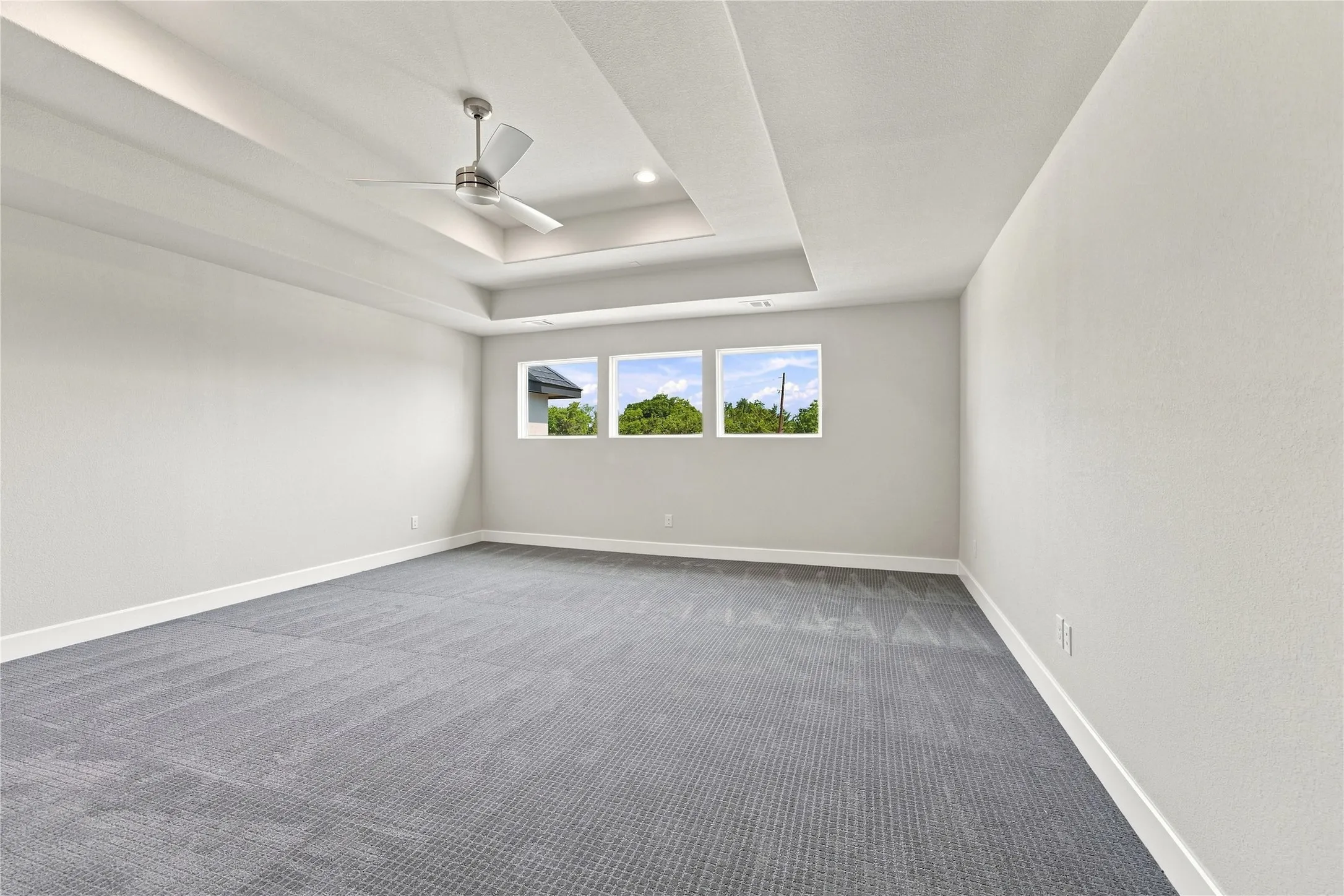 Spacious room featuring a tray ceiling with recessed lighting, a ceiling fan, plush gray carpeting, and multiple windows