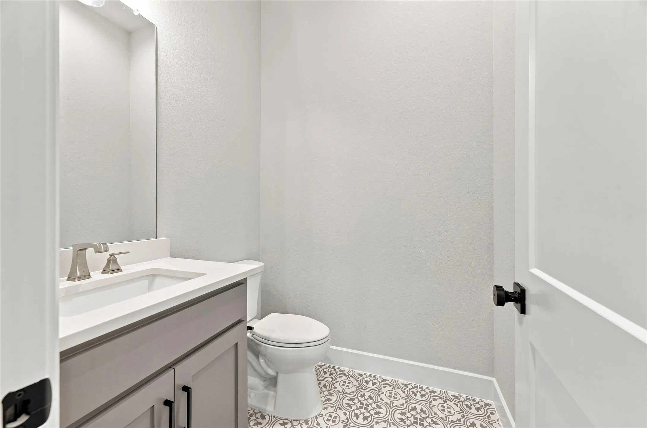 Patterned floor tile, white vanity countertop, undermount sink, brushed nickel faucet, and gray vanity cabinetry
