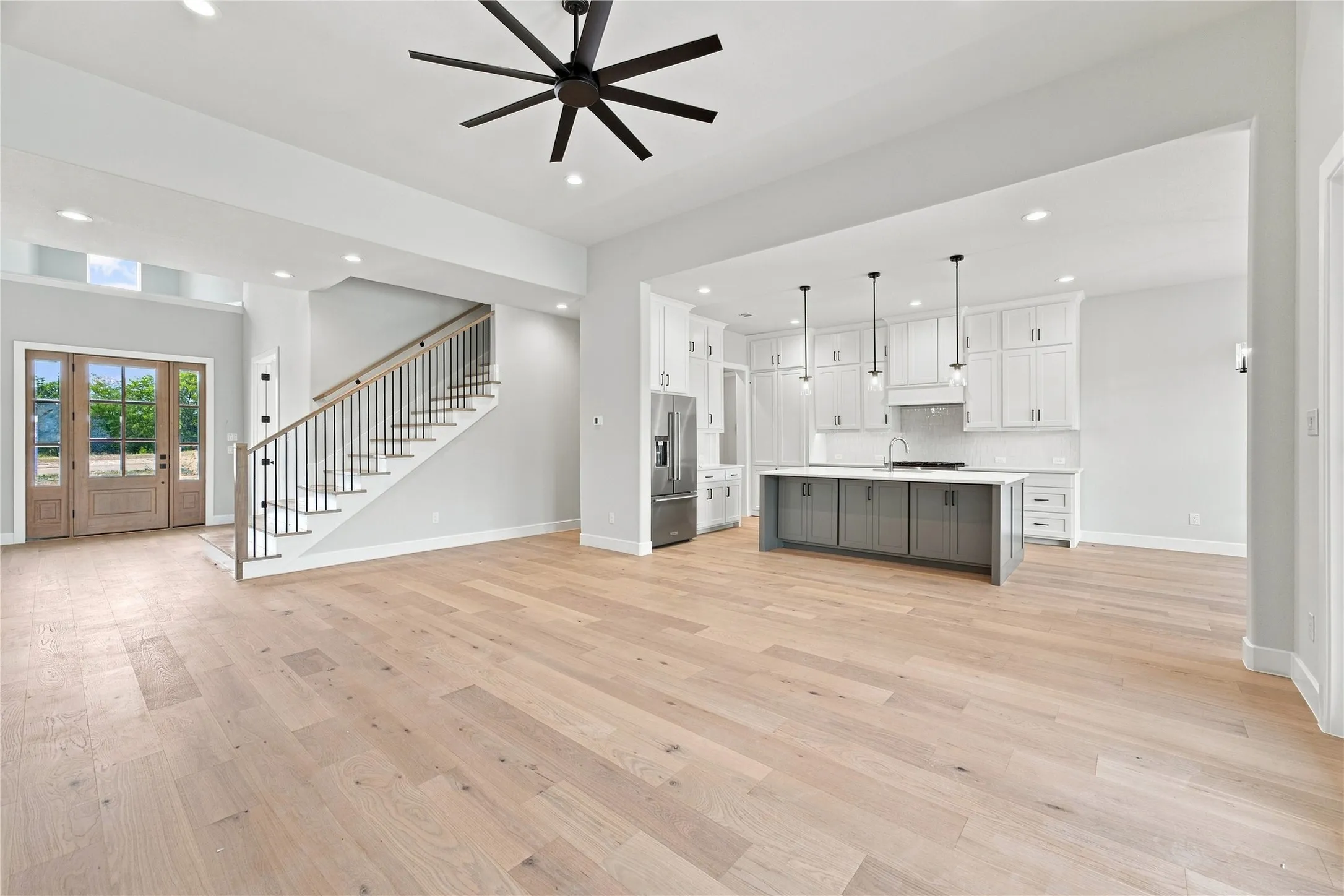Open-concept living area featuring wood-finish flooring, a modern kitchen island with pendant lighting, white cabinetry, a stainless steel refrigerator, and a staircase with metal balusters