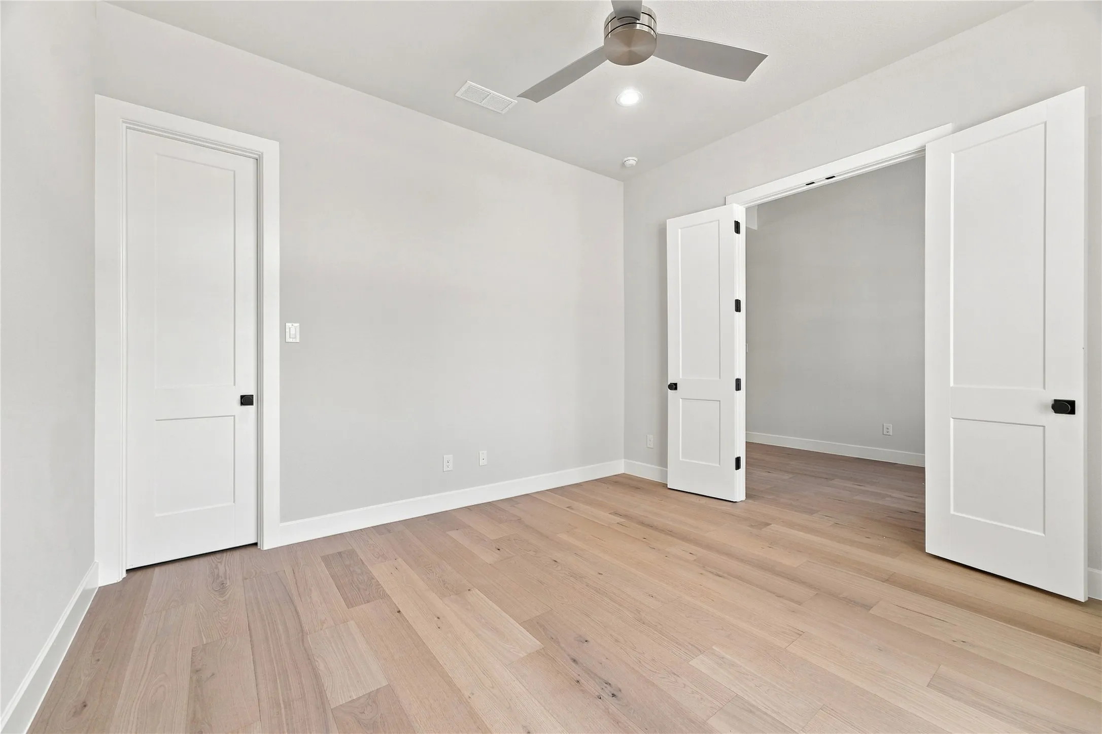 Light wood-finish flooring throughout, bright white walls, contemporary ceiling fan with integrated lighting, recessed ceiling light, and white paneled doors with matte black hardware