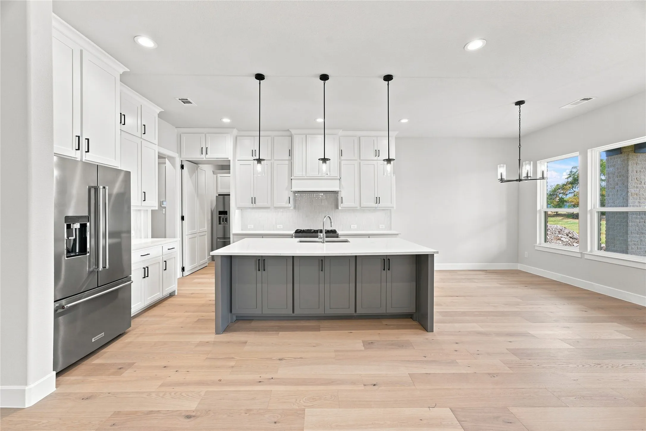 Kitchen featuring a large island with gray cabinetry, stainless steel appliances, white perimeter cabinetry, light wood-finish flooring, and recessed lighting. Butler pantry open.