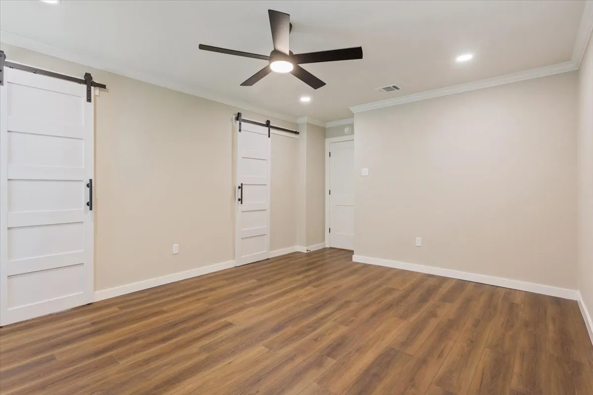 Spacious primary bedroom featuring LVP wood-finish flooring, barn-door access to the ensuite bathroom, a ceiling fan with integrated lighting, and recessed ceiling lights
