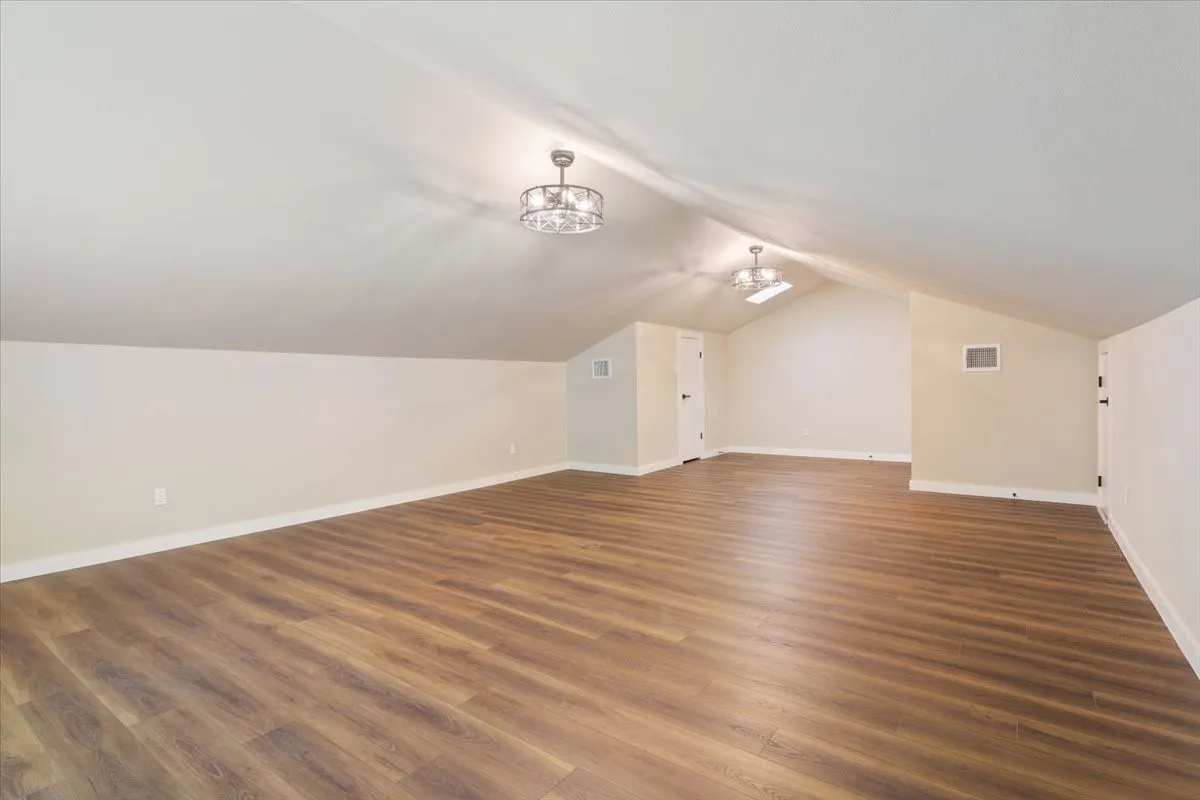 Spacious 4th bedroom / bonus room featuring LVP wood-finish flooring, a vaulted ceiling, two ceiling-mounted decorative light fixtures with ceiling fans (mini), and attic access