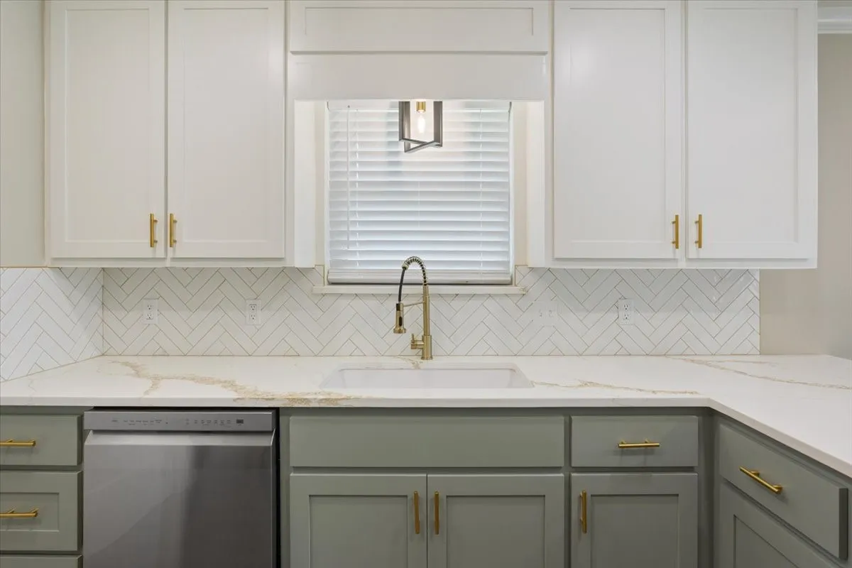 Kitchen featuring a large farm style sink with gooseneck faucet, quartz countertops, white herringbone tile backsplash, and two-tone cabinetry with gold-finish hardware