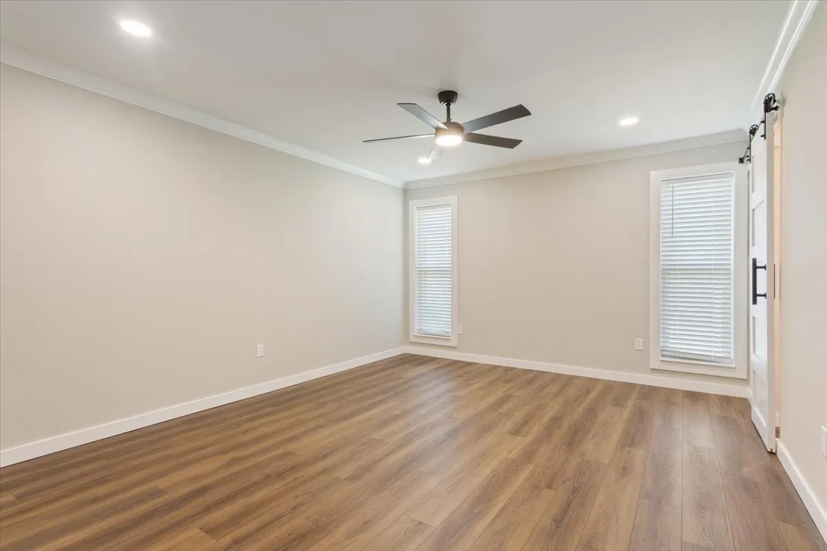 Spacious primary bedroom featuring LVP wood-finish flooring, barn-door access to the ensuite bathroom,two large windows with blinds, a ceiling fan with integrated lighting, and recessed ceiling lights