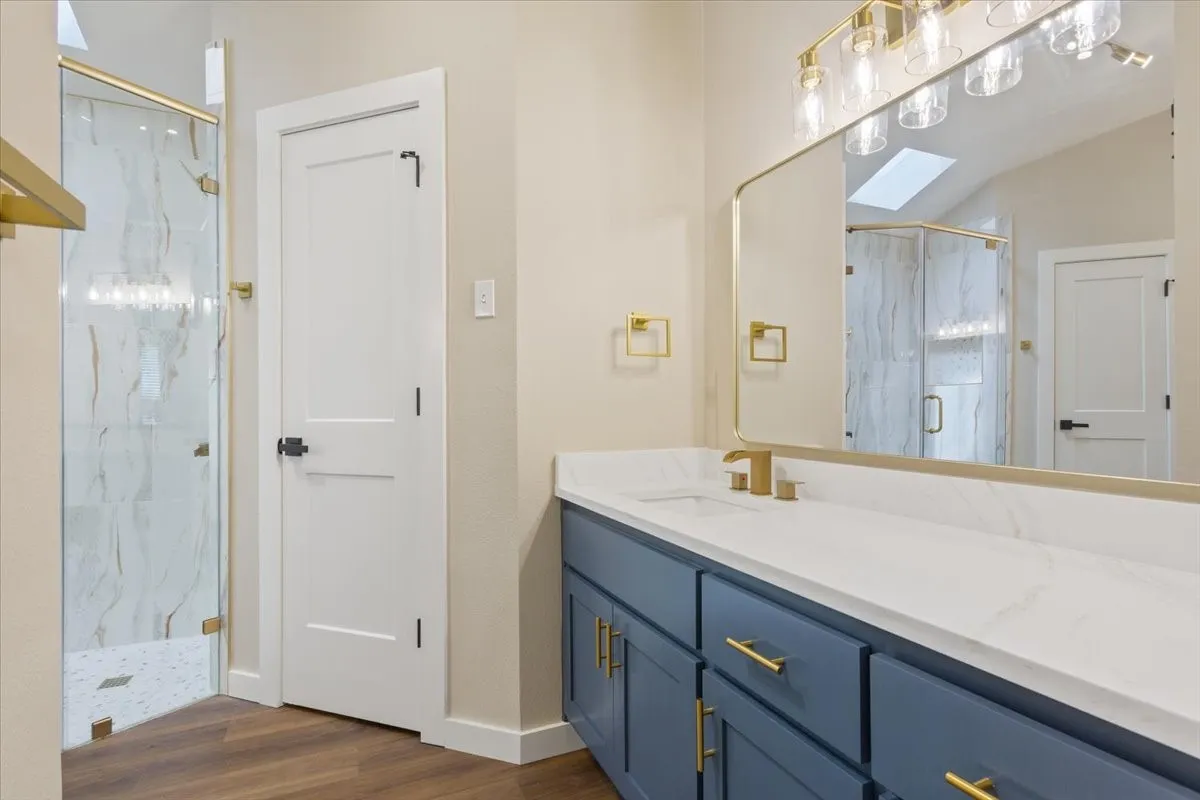 Primary ensuite bathroom featuring a large walk-in shower with custom glass, spacious vanity with blue cabinetry, gold-tone hardware, and a white quartz countertop