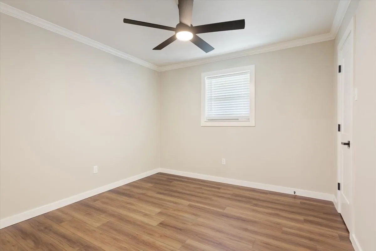 Spacious room featuring wood-finish flooring, a contemporary ceiling fan with integrated lighting, a white framed window with blinds, and clean crown molding