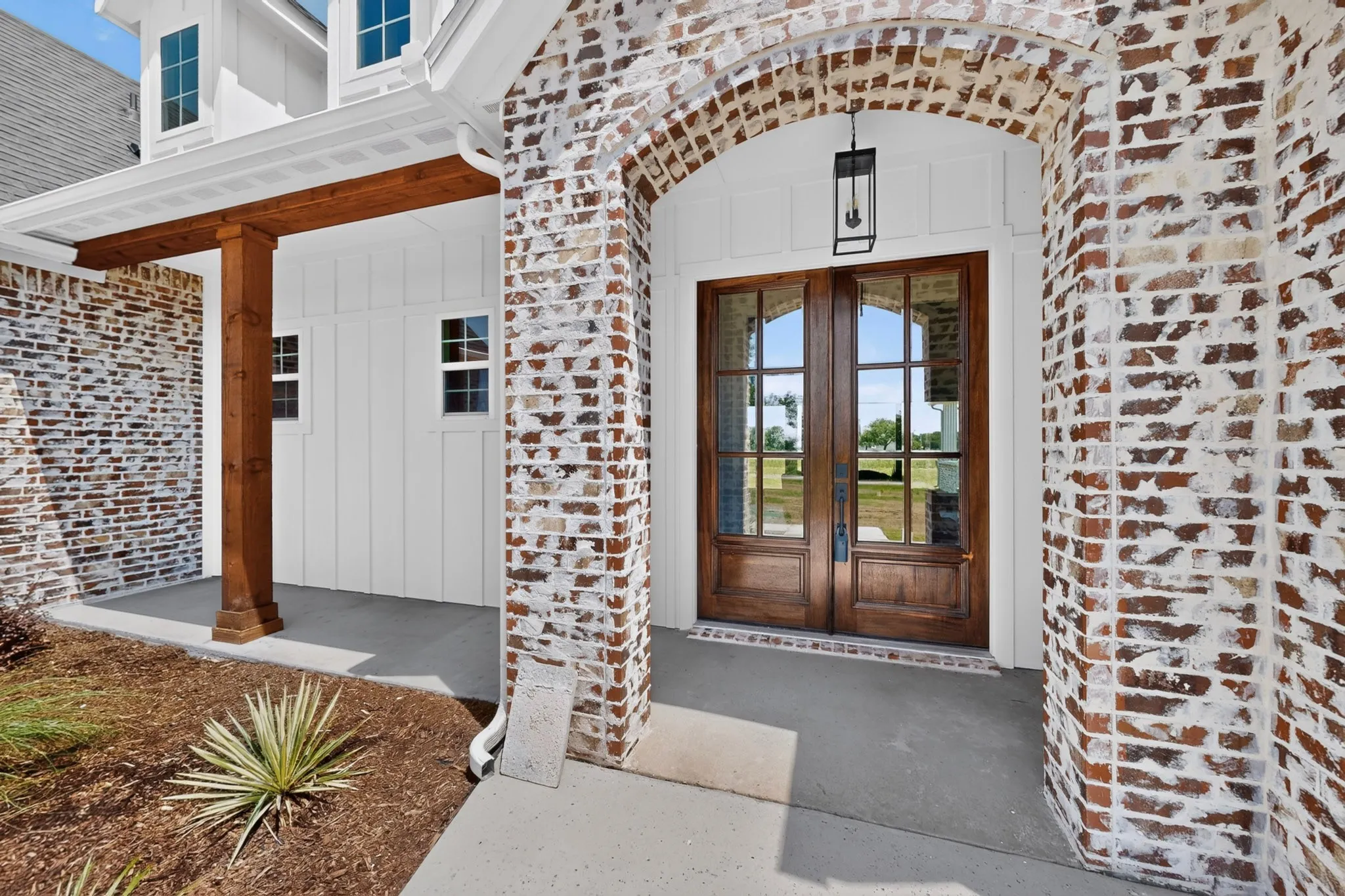 Brick archway entrance featuring double wood-finish doors with glass panels, white vertical plank siding, a wood-beam porch column, and a modern exterior light fixture