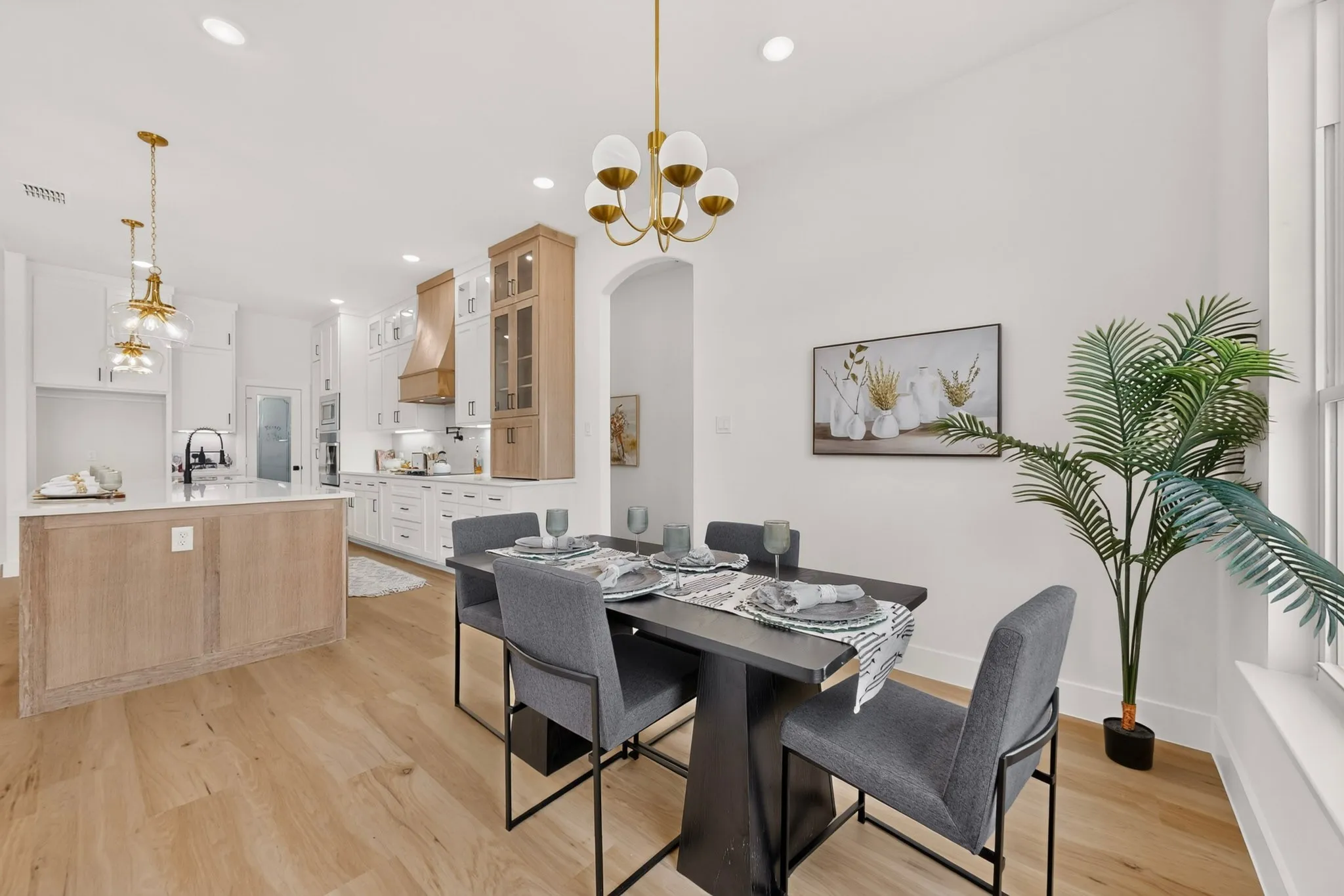 Open-concept living space featuring wood-finish flooring, a contemporary kitchen with white and natural wood cabinetry, a central island with a light-colored countertop, and a dining area illuminated by a modern chandelier