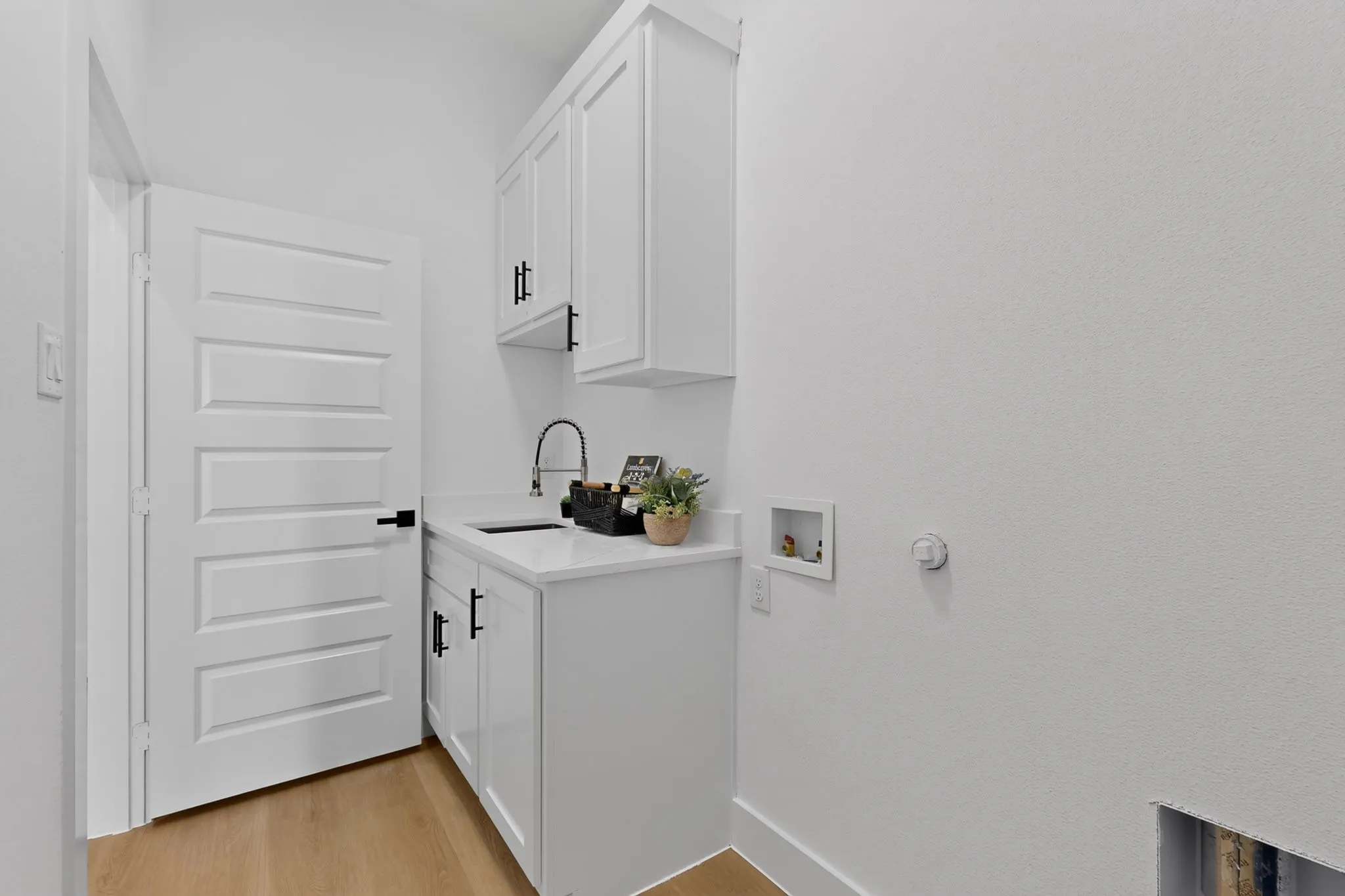 Utility room featuring light wood-finish flooring, white paneled cabinetry with dark hardware, a white countertop, and a dark basin sink with a gooseneck faucet