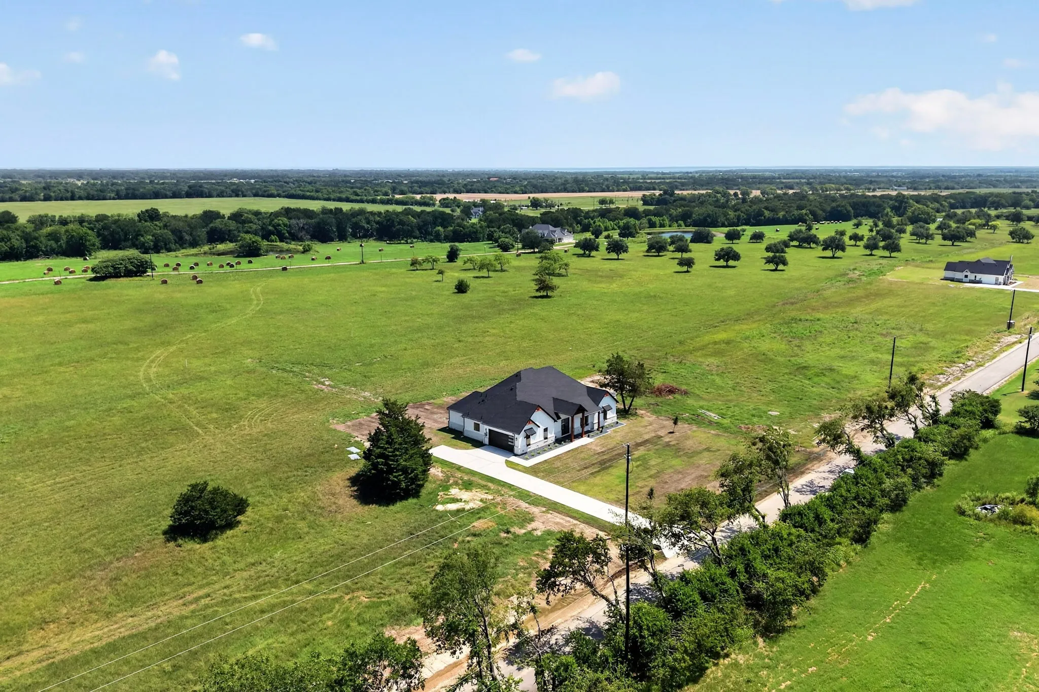 Expansive green landscape featuring a single-story residence with a dark roof, surrounded by mature trees, a private driveway, and a distant paved road