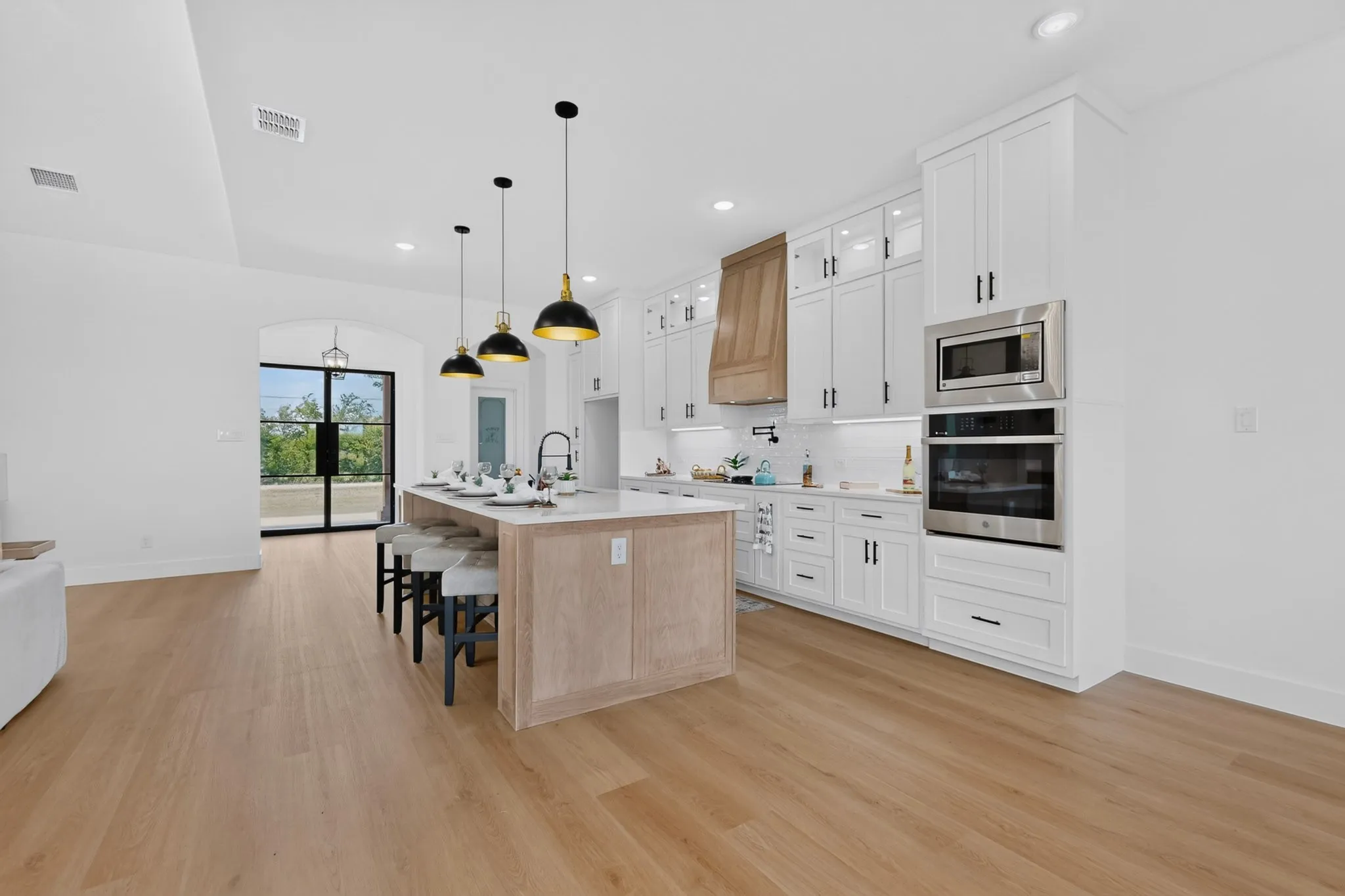 Spacious open-concept kitchen featuring a large center island, white cabinetry, integrated stainless steel appliances, a wood-finish range hood, and light wood-finish flooring
