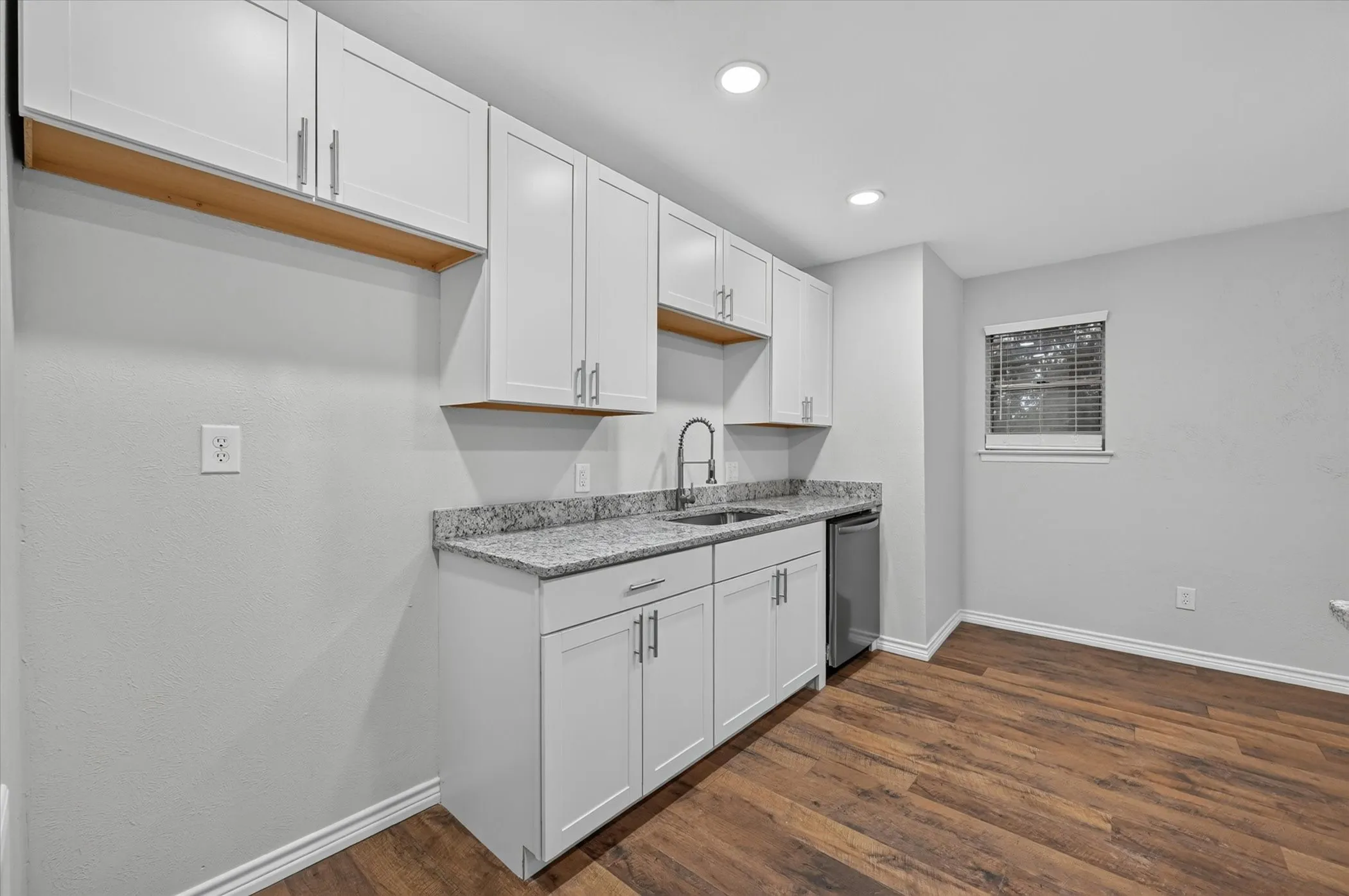 Kitchenette featuring white shaker cabinetry, granite countertops, a stainless steel sink with a gooseneck faucet, a compact refrigerator, and wood-finish flooring