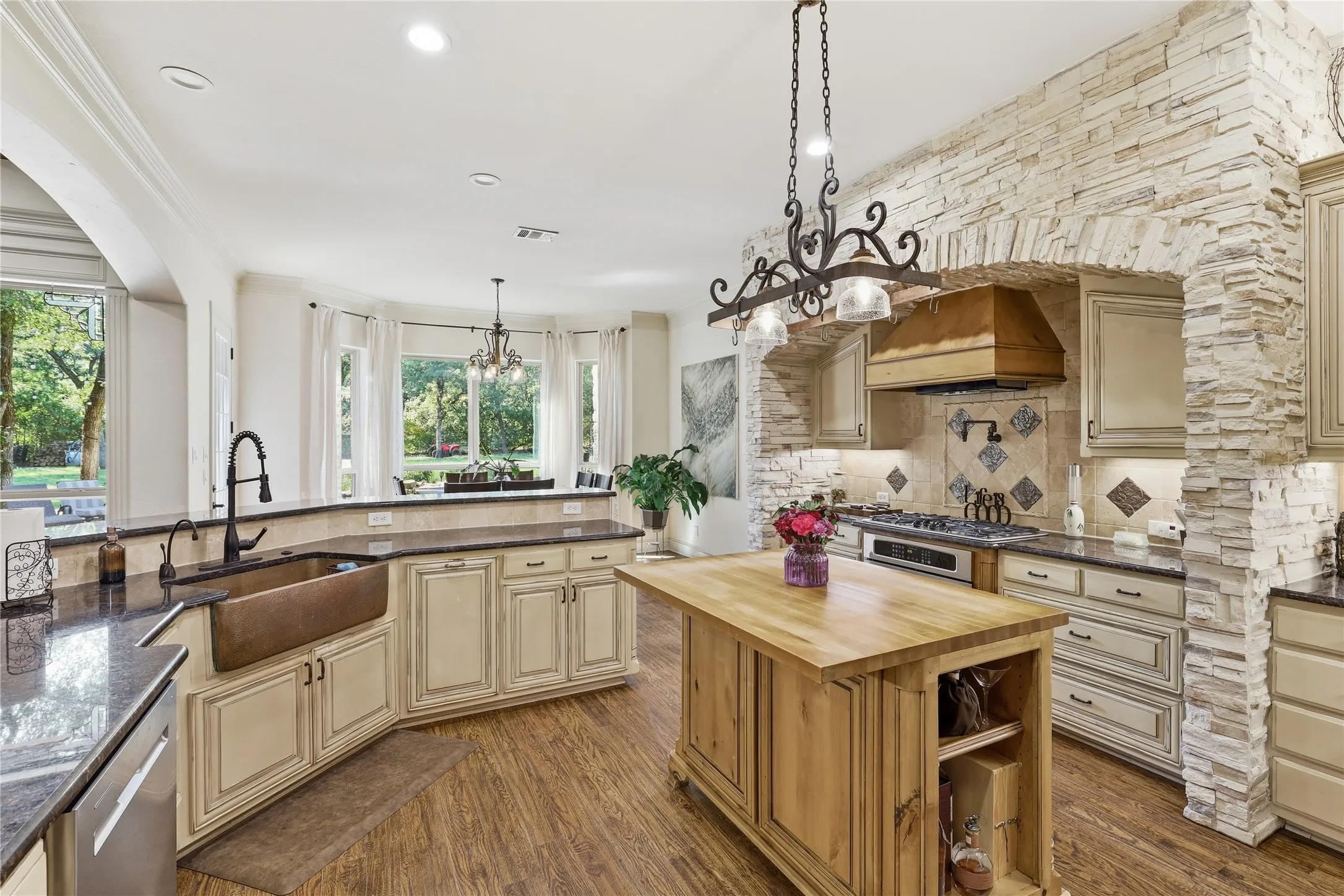 Gourmet kitchen featuring a stone-clad range hood, custom cabinetry, granite countertops, a copper farmhouse sink, and a portable butcher block island