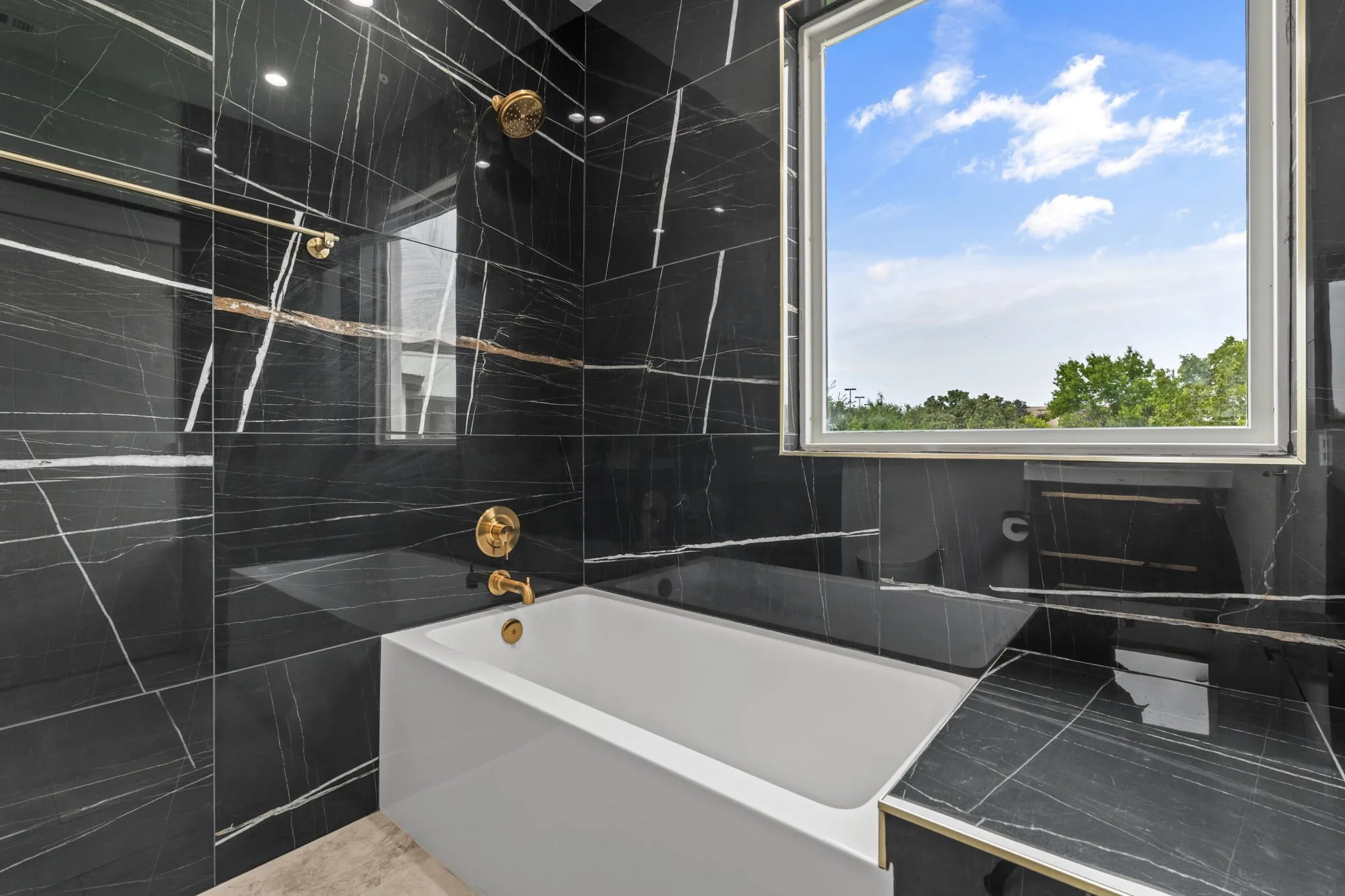 Guest bathroom featuring a deep soaking tub, black marble-look wall tiles with white and gold veining, polished gold fixtures, and a large window with white framing