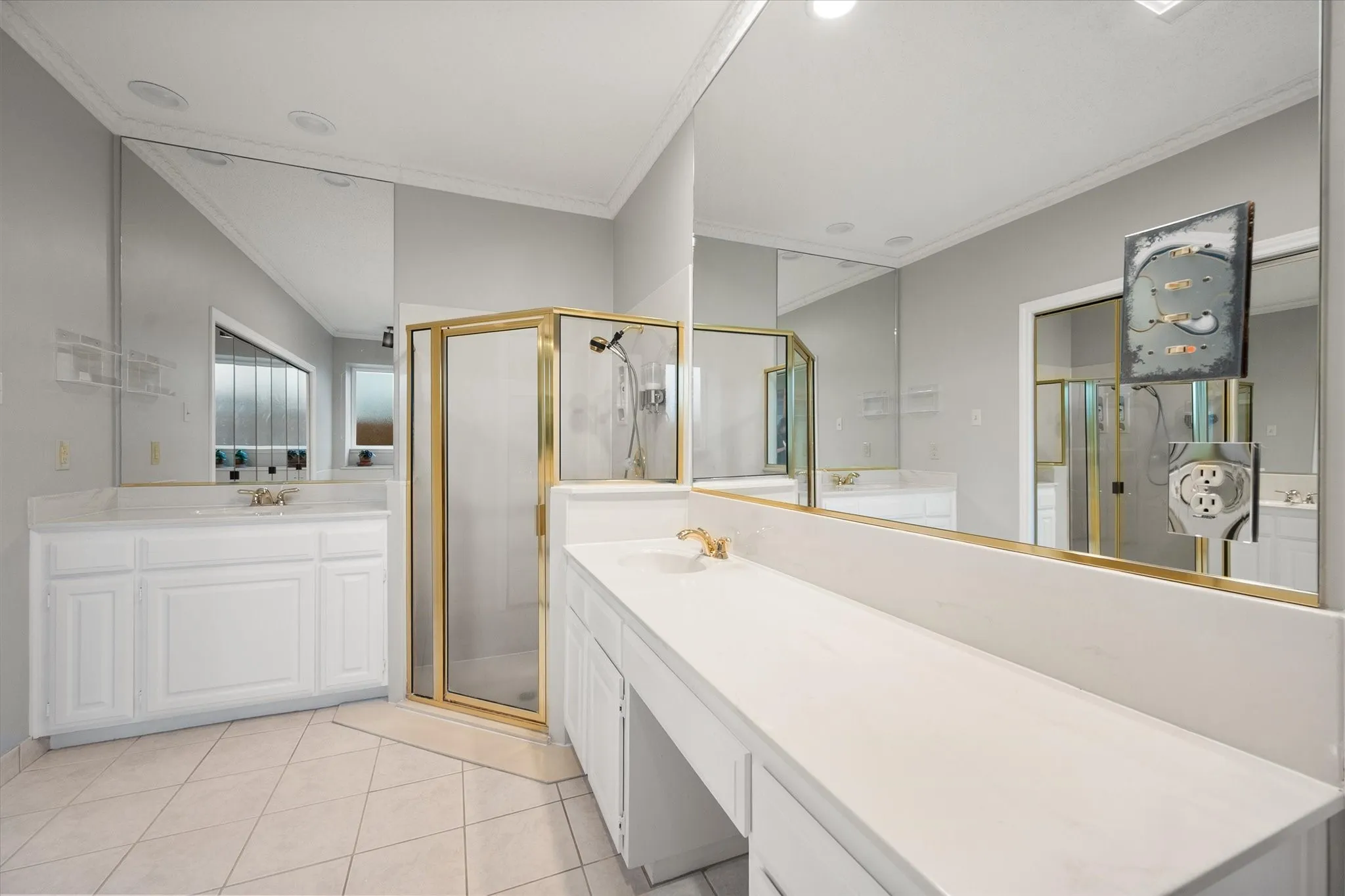 Spacious bathroom featuring a glass-enclosed shower with gold-tone framing, extensive vanity countertop space, dual white cabinetry, large wall-mounted mirrors, and tiled flooring