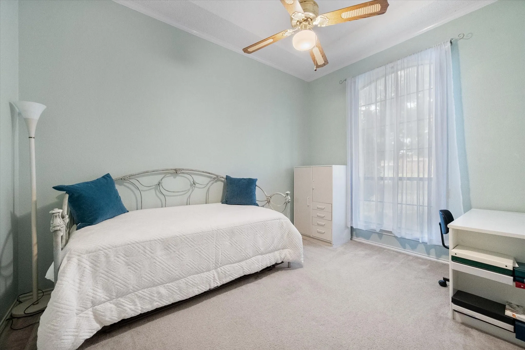Carpeted interior with light-toned walls, featuring a wide window with sheer curtains, a ceiling fan with light fixture, and crown molding