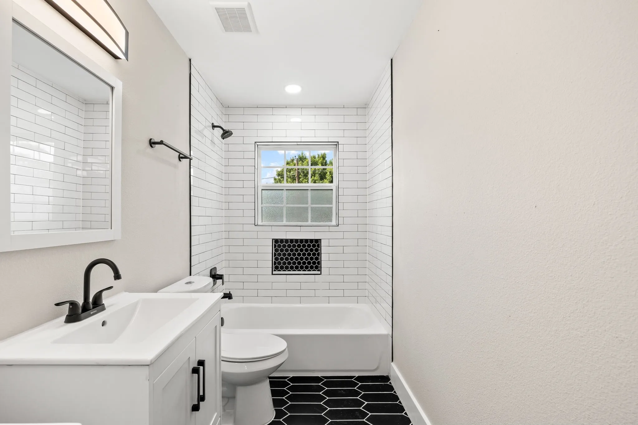Bathroom featuring a white vanity with matte black hardware and faucet, a white subway tile shower surround with black grout, a built-in shower niche with black hexagonal tile, black hexagonal floor tile, and a single window