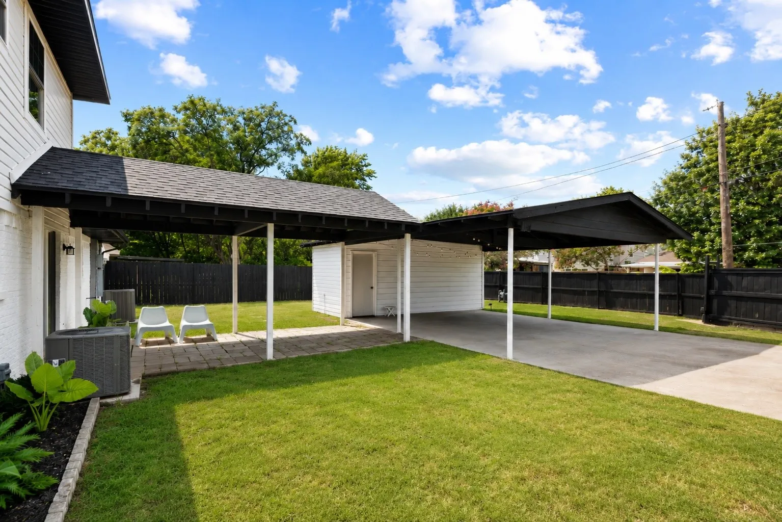 Expansive backyard featuring a manicured lawn, a covered patio with paver stones, an attached carport with concrete flooring, a detached storage shed, and a privacy fence