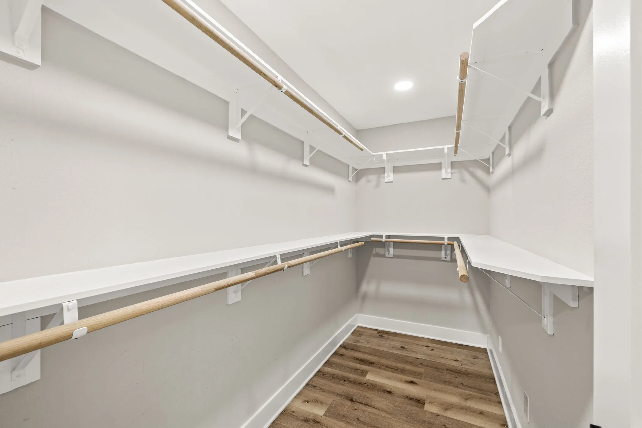 Walk-in closet featuring light gray walls, wood-finish flooring, white baseboards, and built-in white shelving with natural wood hanging rods