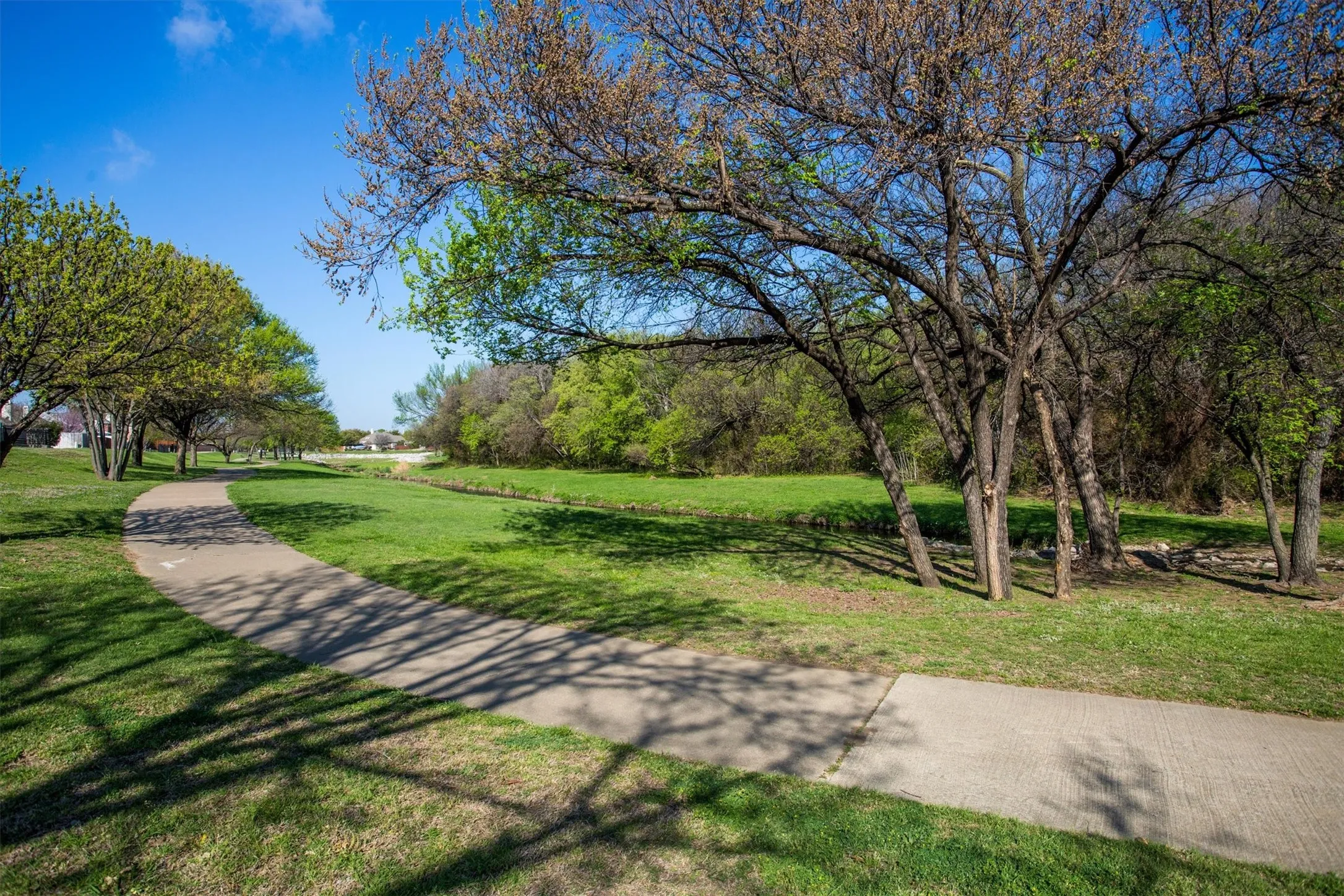 Paved pathway bordered by manicured lawn and mature trees, featuring a natural creek bed and distant residential structures