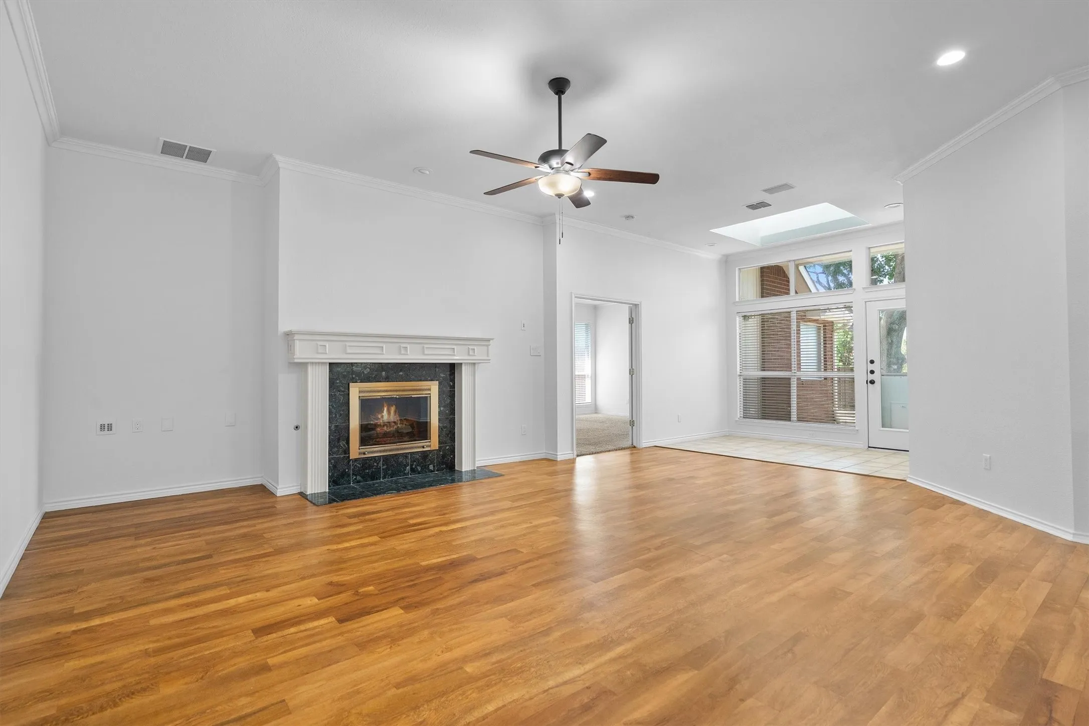 Spacious room featuring wood-finish flooring, a fireplace with a dark stone surround and white mantel, a ceiling fan, a skylight, and large glass doors