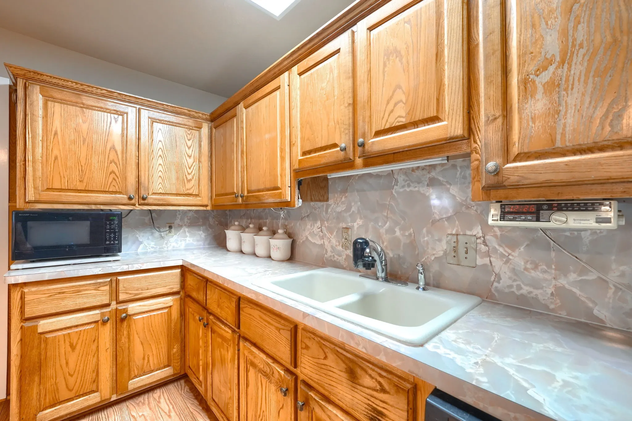 Corner kitchen counter featuring a double basin sink, light wood-finish cabinetry, and a patterned backsplash
