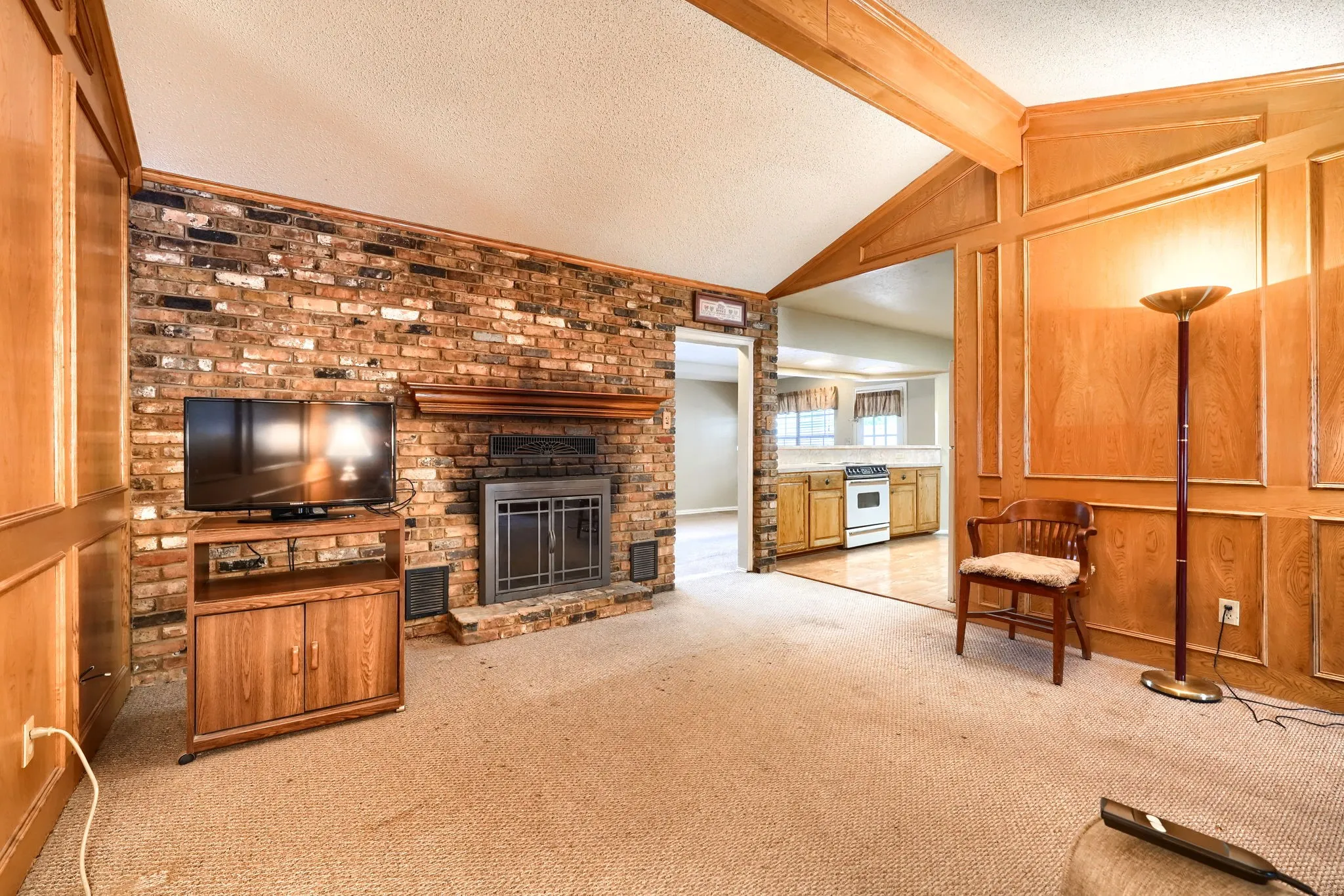 Carpeted living area featuring a brick fireplace with a wood mantel, exposed wood beams, and paneled walls