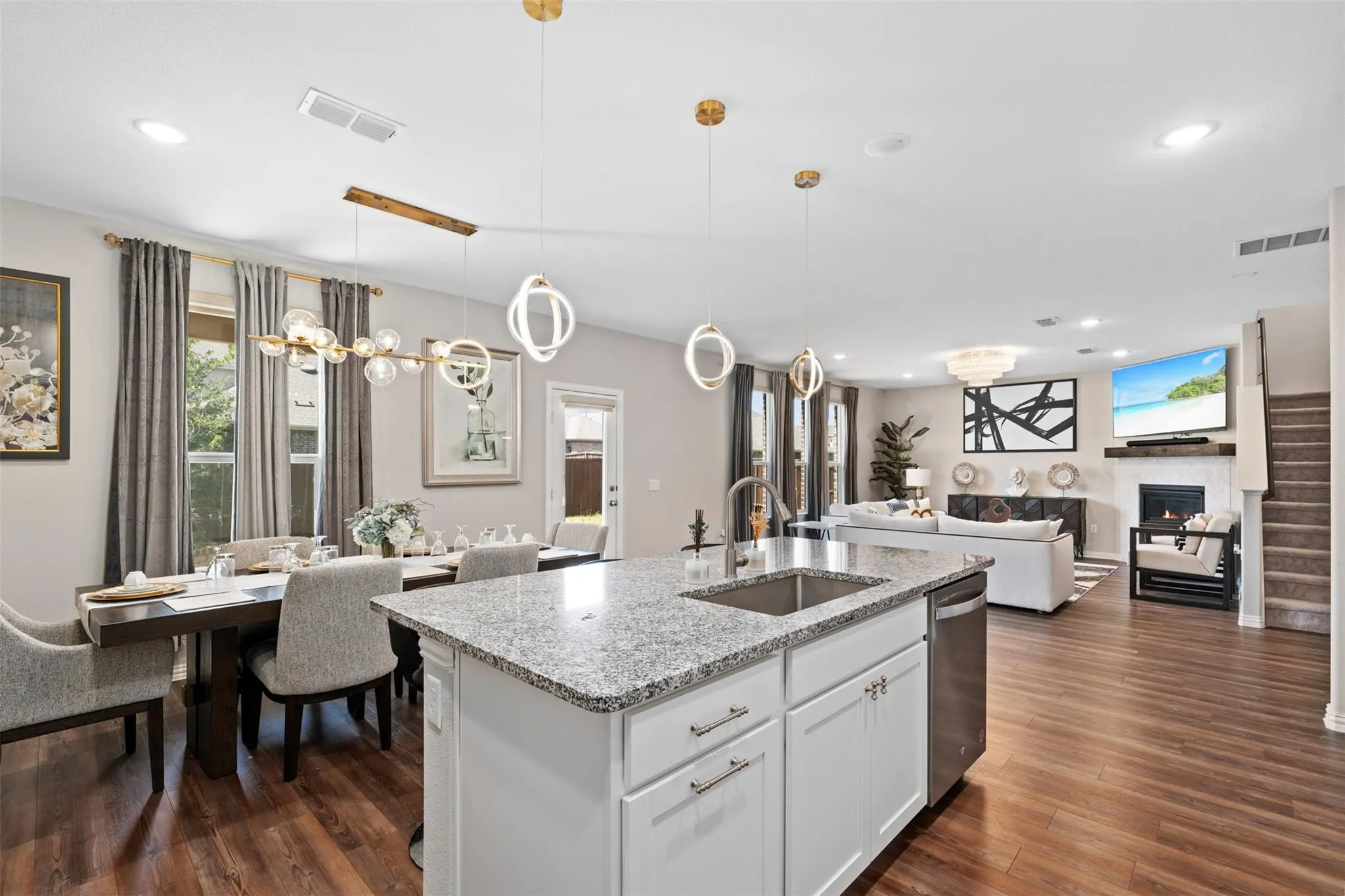Open-concept living area featuring wood-finish flooring, a kitchen island with stone countertops, a dining area with contemporary lighting, a living room with a fireplace, and a staircase