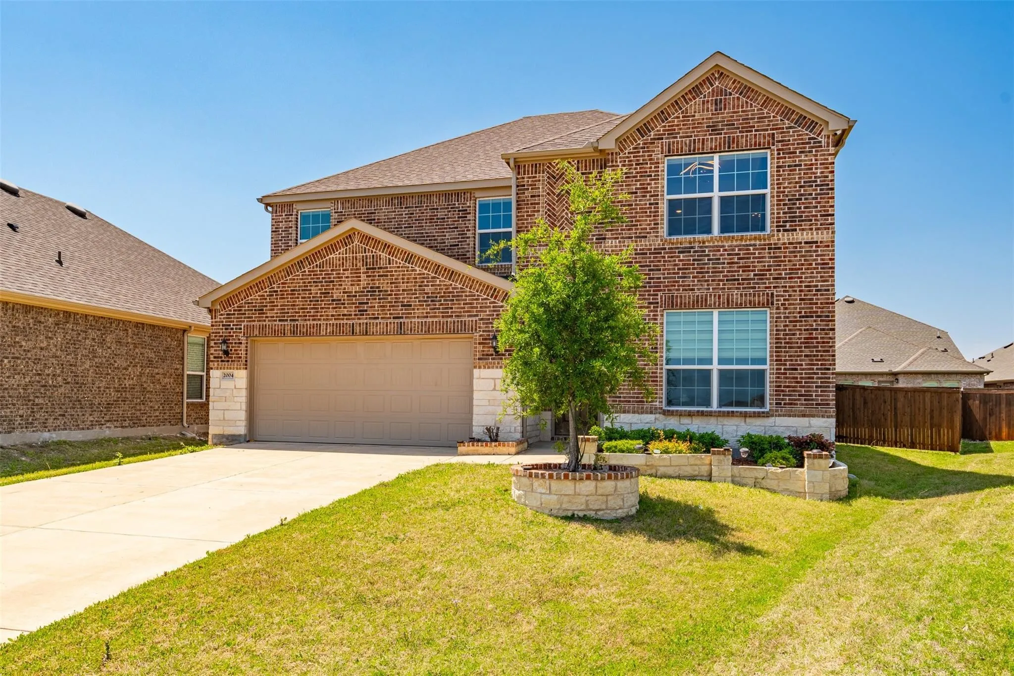 Two-story brick exterior with stone accents, a two-car garage, concrete driveway, and front lawn landscaping featuring a planted tree and stone-lined garden beds