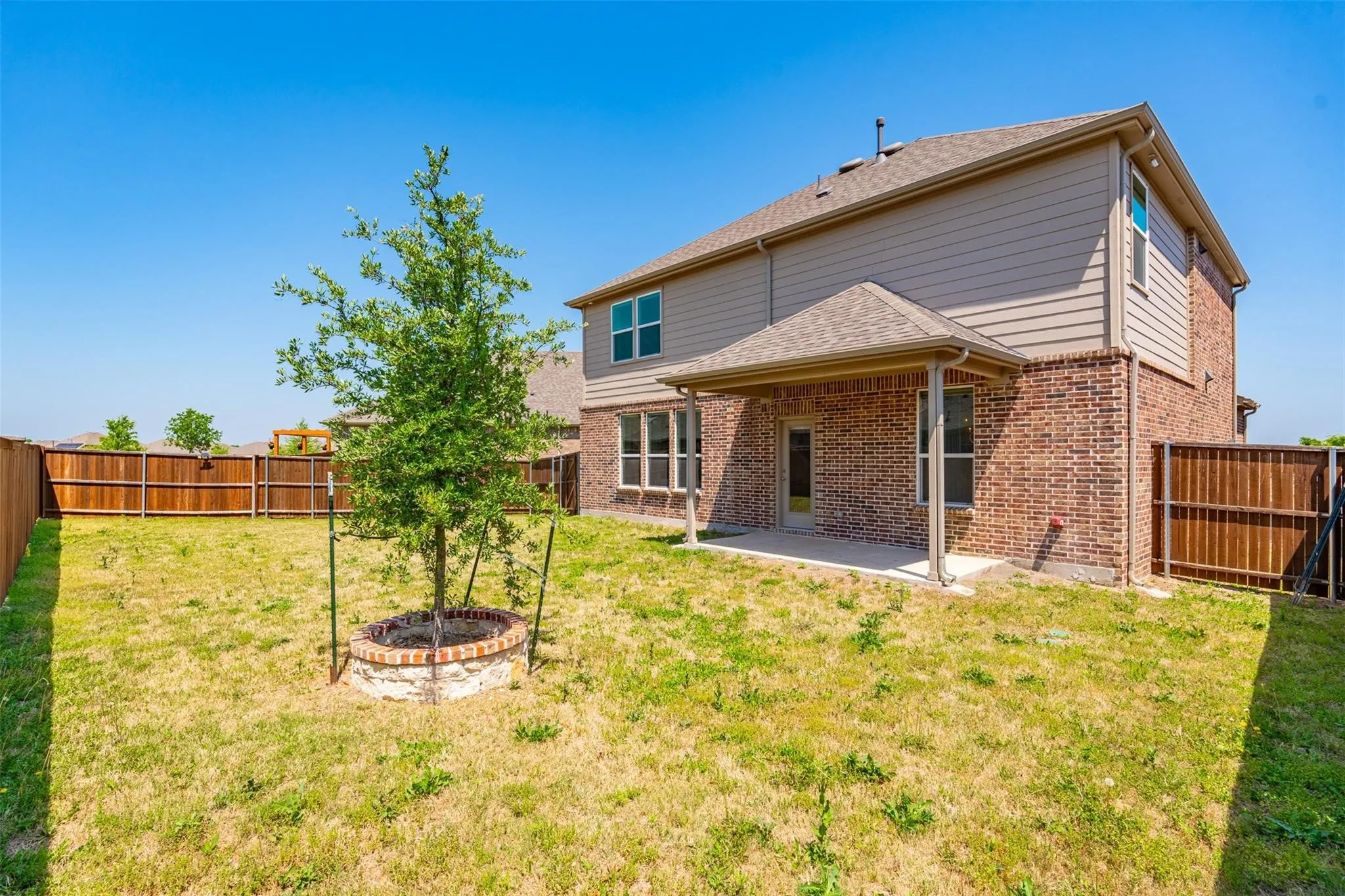 Spacious backyard featuring a covered patio, brick and siding exterior, fenced perimeter, and a young tree with brick planter