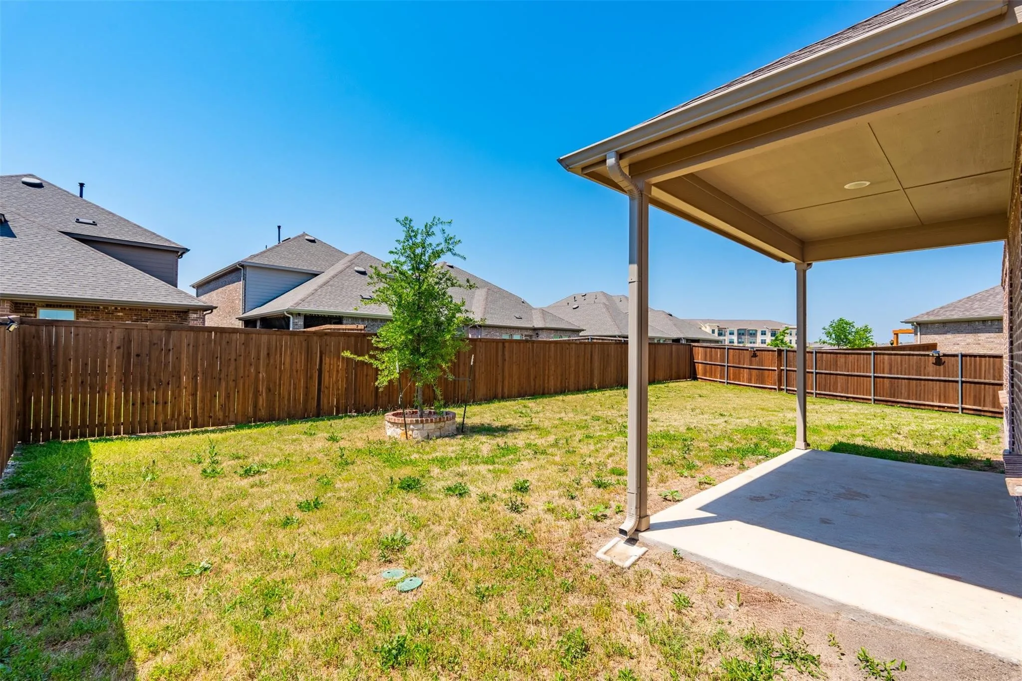 Expansive backyard featuring a covered patio, full perimeter wood fencing, and a young tree with a stone-edged planting area