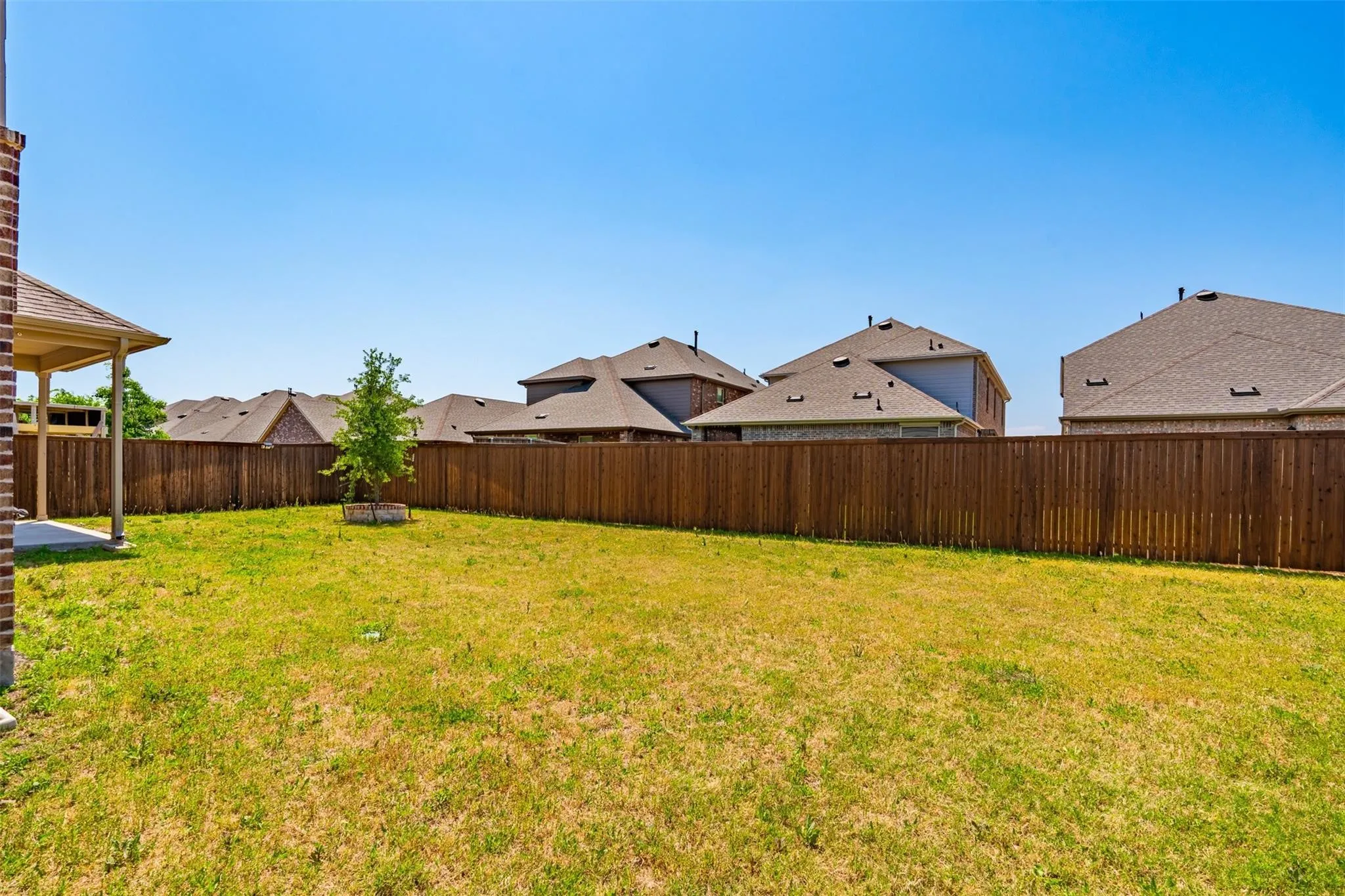 Expansive backyard featuring a privacy fence, mature sapling, and a covered patio with a concrete slab and brick pillar
