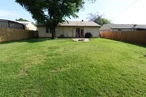 Spacious rear yard featuring a large grass area, mature shade tree, full privacy fencing, and a concrete patio with exterior access