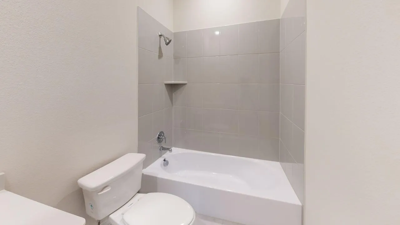 Full bathtub with chrome fixtures, light gray tile surround, and an integrated corner shelf