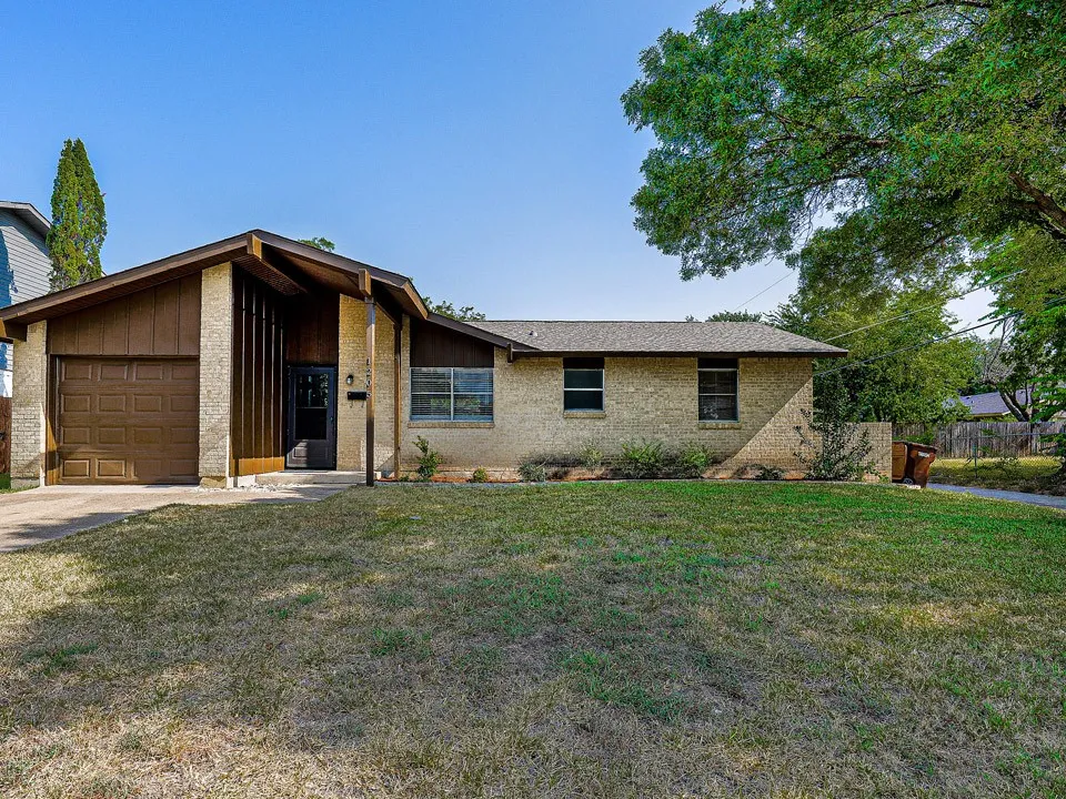 Brick ranch-style home featuring a single-car attached garage with wood-finish door, manicured front lawn, and mature landscaping including established trees and evergreen plantings