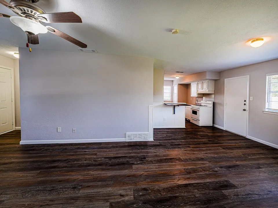 Open living area featuring dark wood-finish flooring, ceiling fan with integrated lighting, and seamless access to a kitchen with white cabinetry and stainless steel appliances