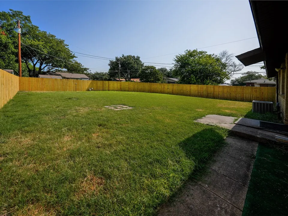 Expansive backyard featuring a manicured lawn enclosed by wood-finish fencing, mature shade trees, concrete patio area, and direct access from the home's rear exit