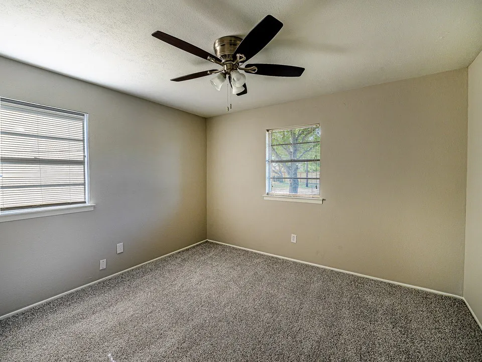 Spacious bedroom showcasing neutral wall finishes, plush carpeting, ceiling fan with integrated lighting, and dual windows providing natural light