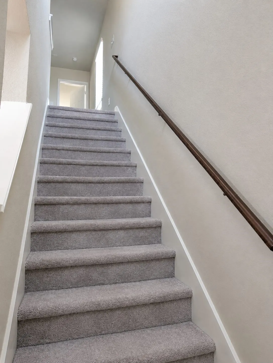 Staircase featuring grey carpeting, white risers, a dark wood handrail, and light neutral wall finishes