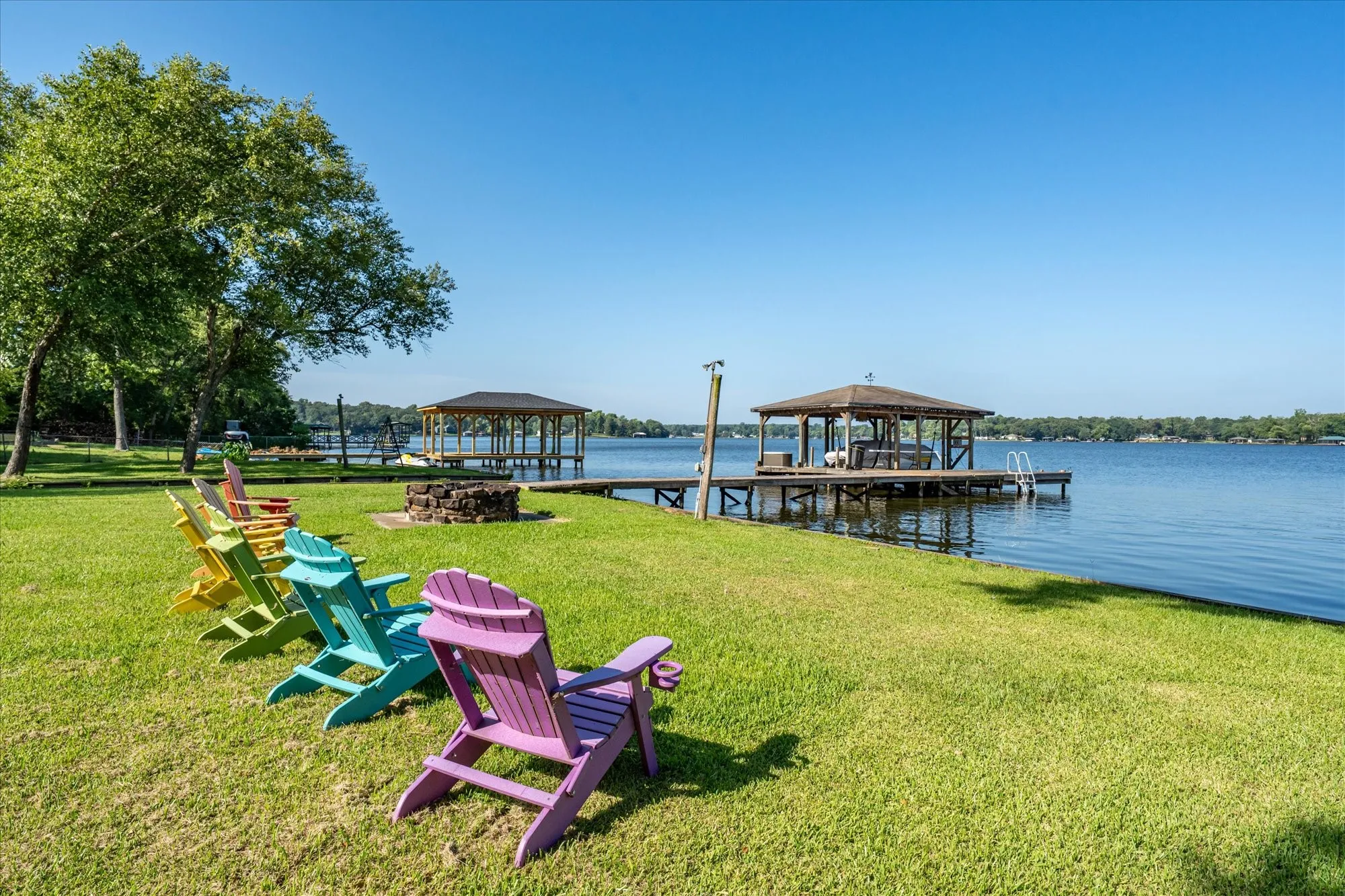 Expansive waterfront property featuring a covered boat dock with a built-in ladder, a shoreline fire pit, and a grassy lawn with mature trees