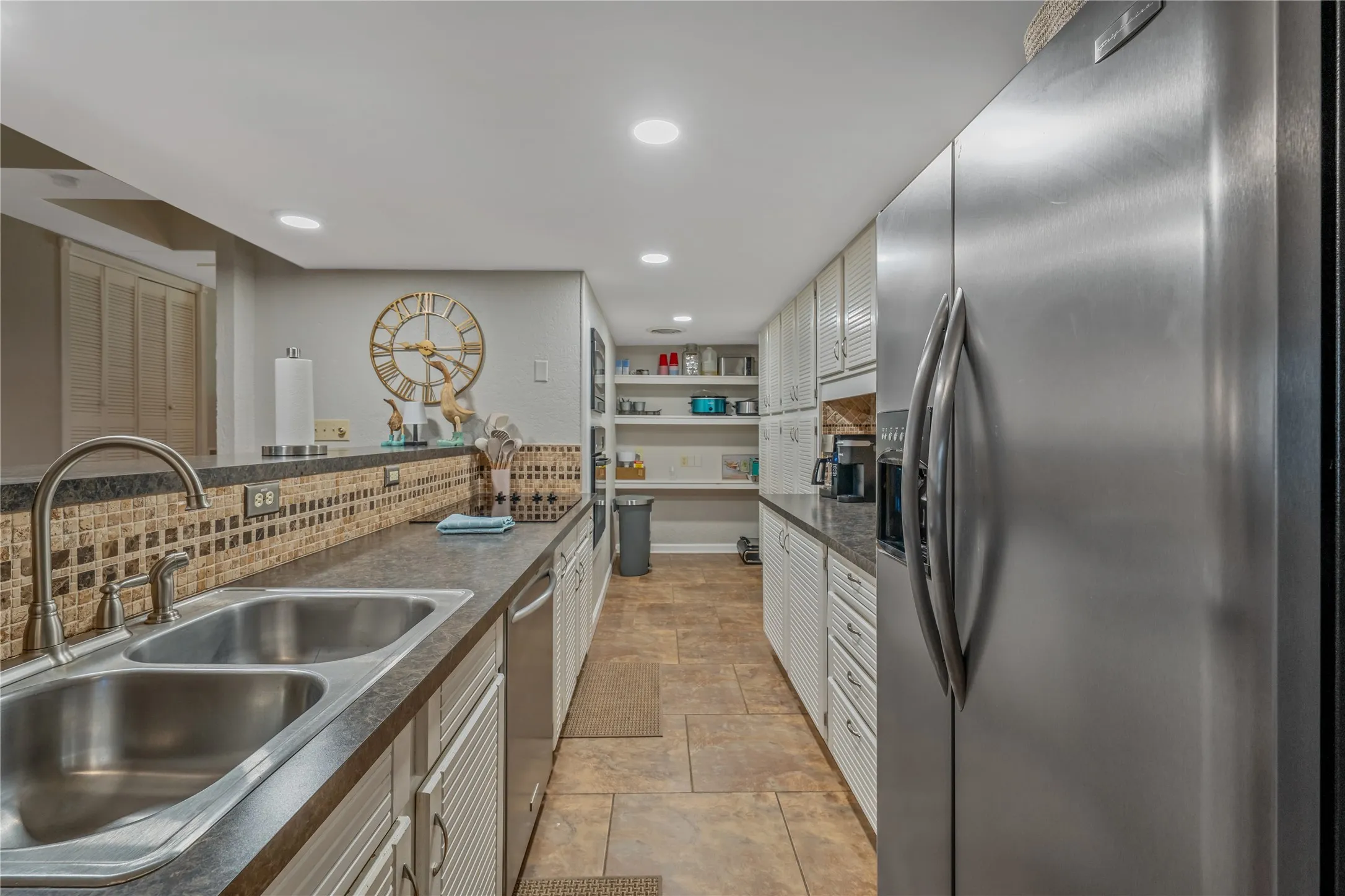 Stainless steel refrigerator, dual basin sink with gooseneck faucet, light-toned tiled flooring, white slatted cabinetry, and recessed lighting