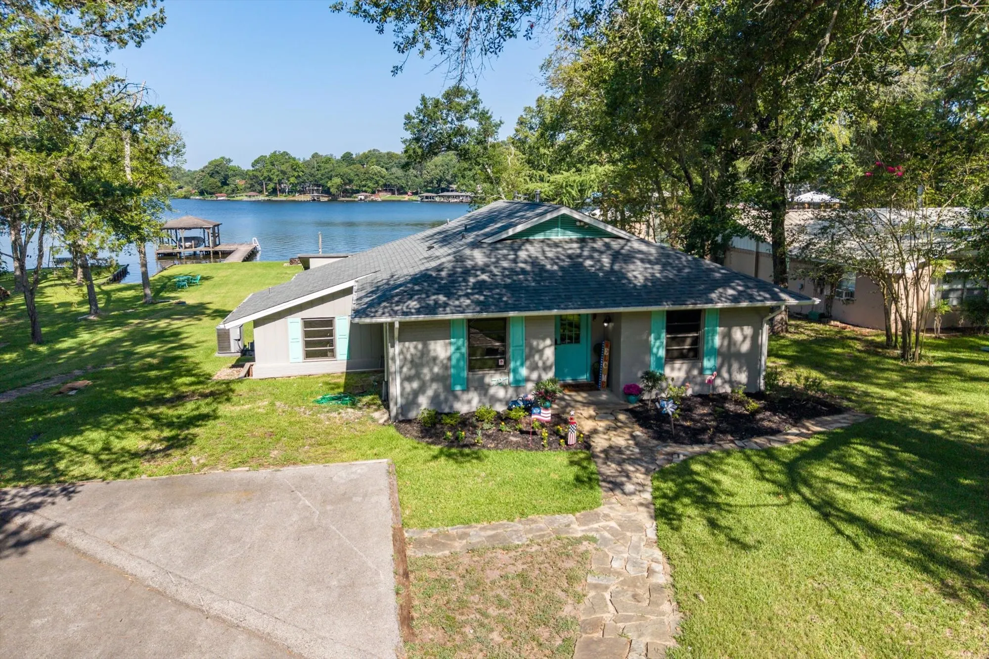Waterfront property featuring a stone walkway, landscaped front beds, a private dock with a covered boat slip, and mature trees