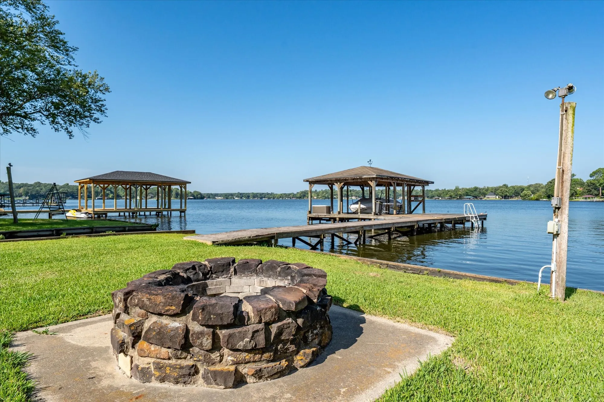 Waterfront property featuring a stone fire pit, two covered boat docks, and a wooden pier with ladder access to the water